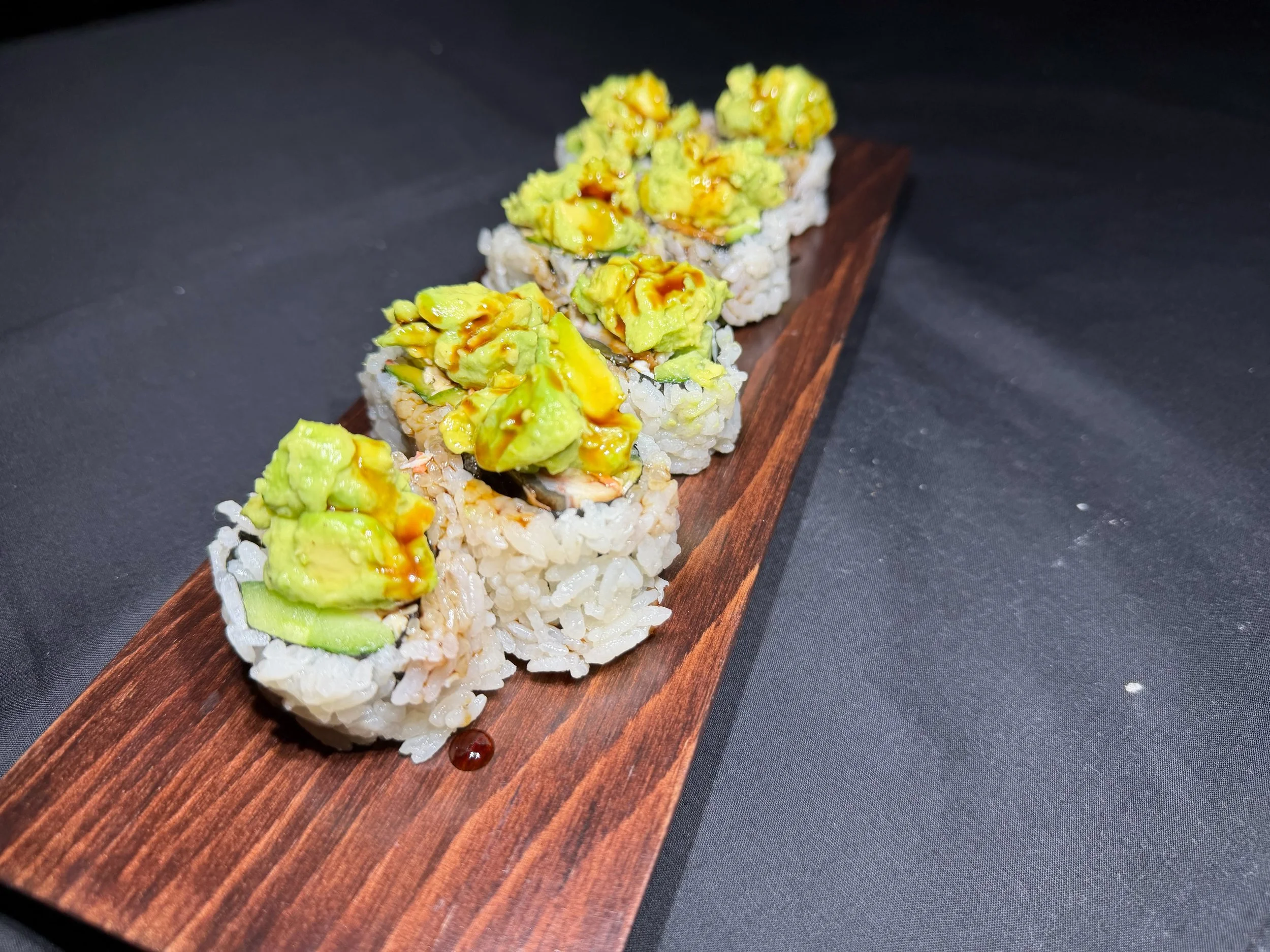 Sushi rolls topped with sliced avocado and drizzled with sauce on a wooden serving board.