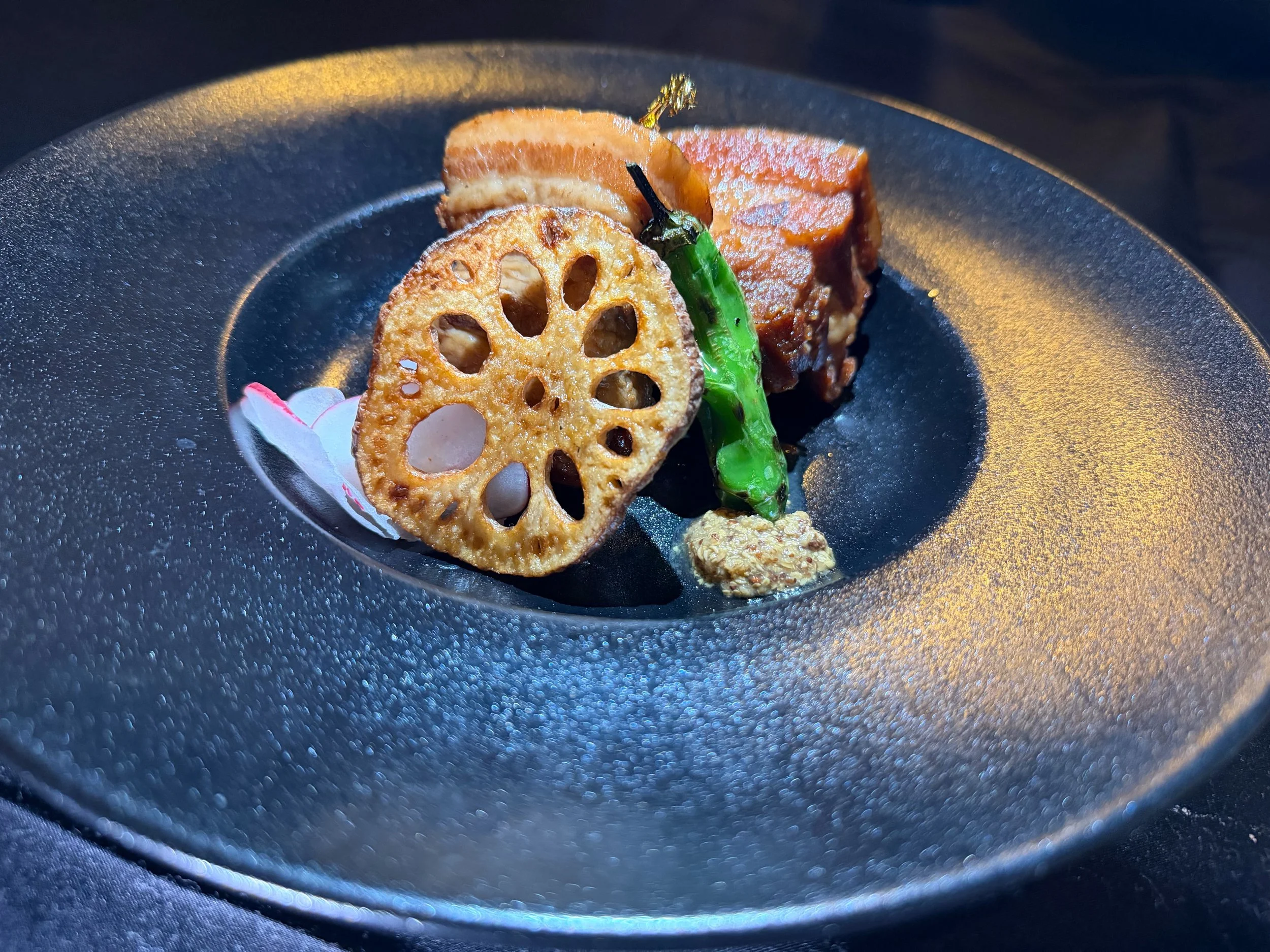 A black textured plate holding a small portion of food: a grilled green chili, a slice of lotus root, a piece of braised meat, a dollop of grainy mustard, and a flower petal garnish.