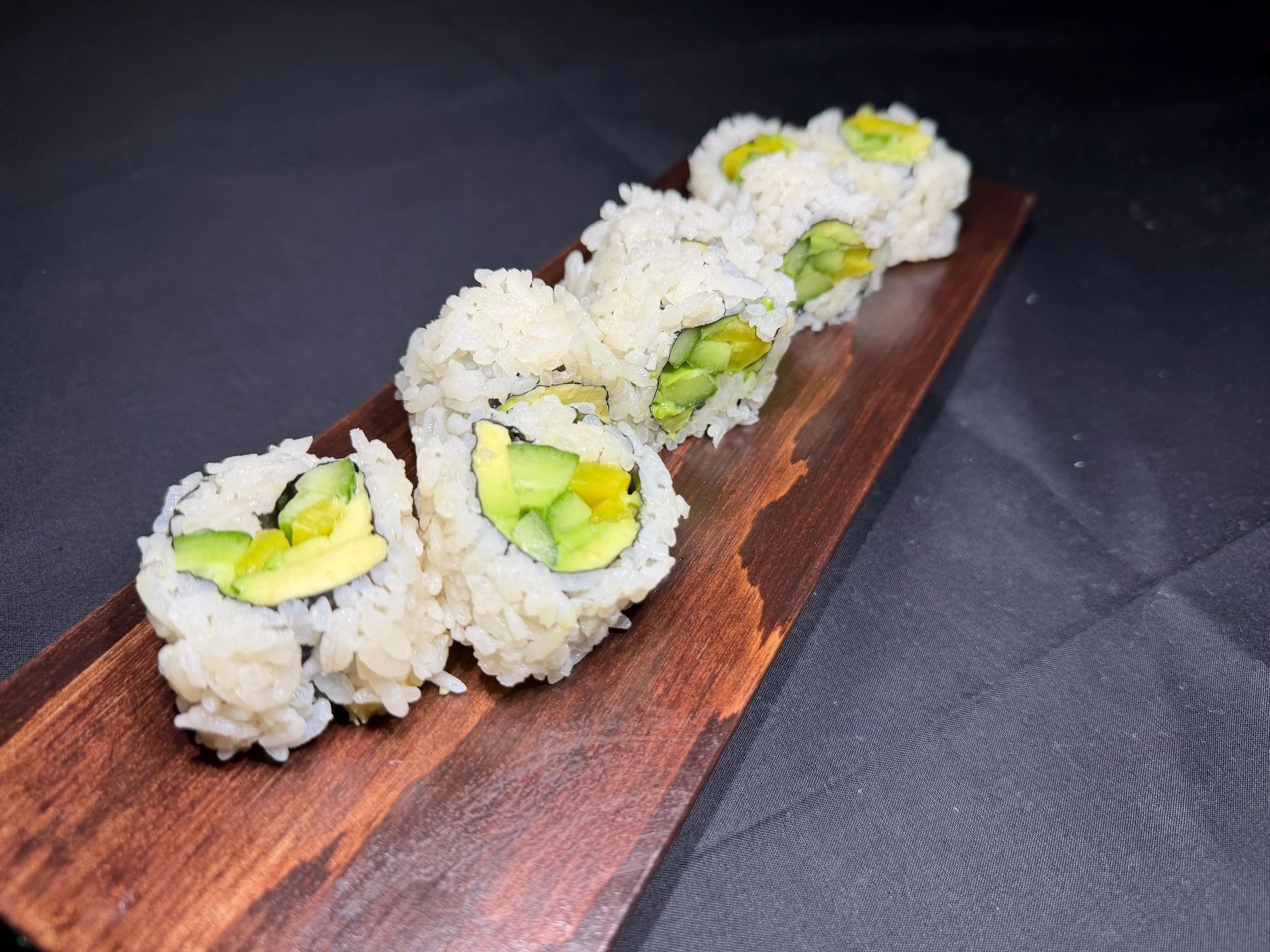 Close-up of six avocado sushi rolls on a long wooden serving board against a black background