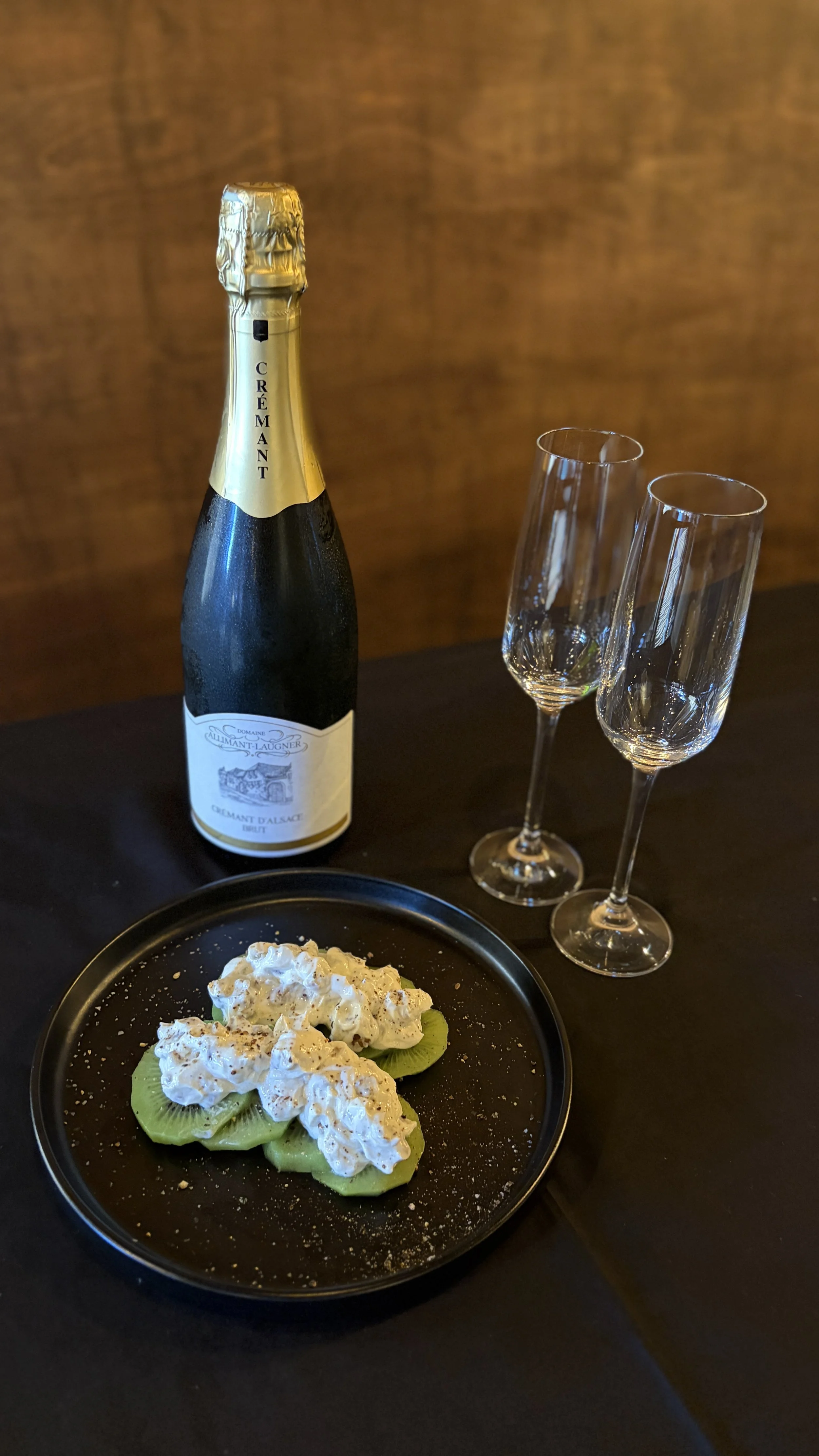 A bottle of sparkling wine, two empty champagne flutes, and a black plate with slices of kiwi topped with whipped cream and crushed nuts on a dark table.