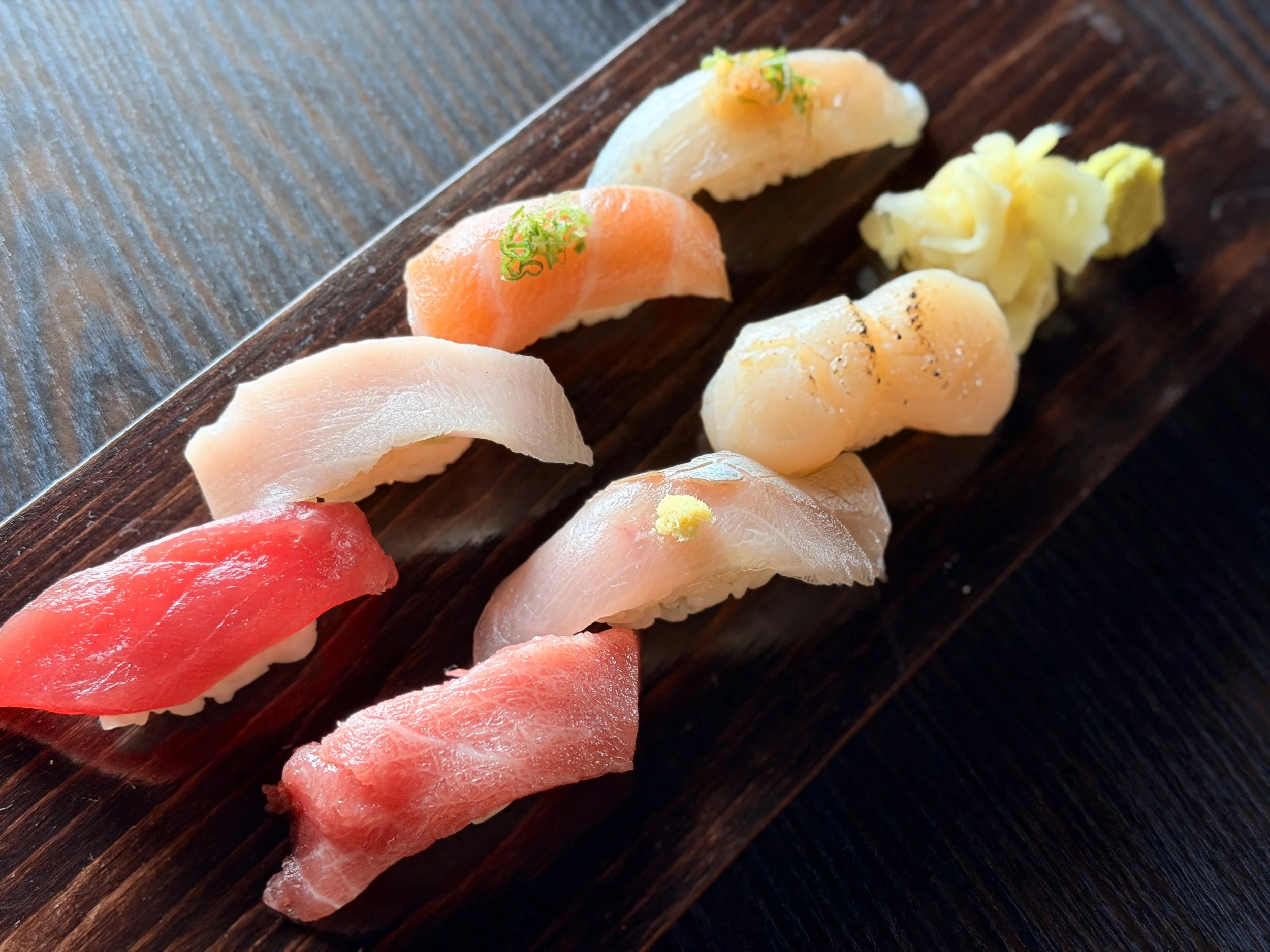 Close-up image of assorted sushi pieces on a dark wooden serving board, including nigiri with salmon, tuna, and white fish, garnished with green herbs.