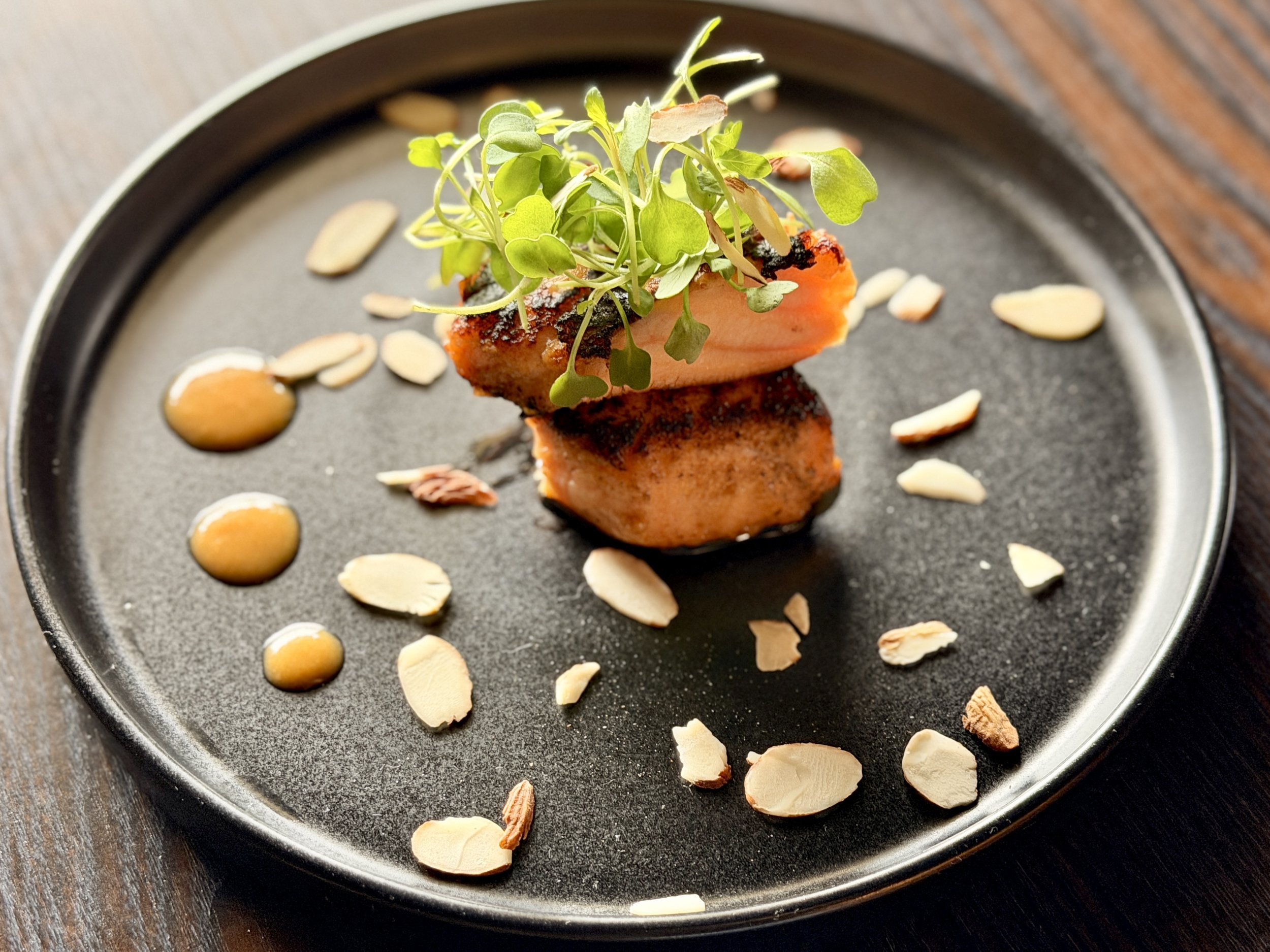 A plated gourmet dish featuring a piece of cooked fish garnished with microgreens, surrounded by almond slices and drizzled with sauce on a dark round plate.
