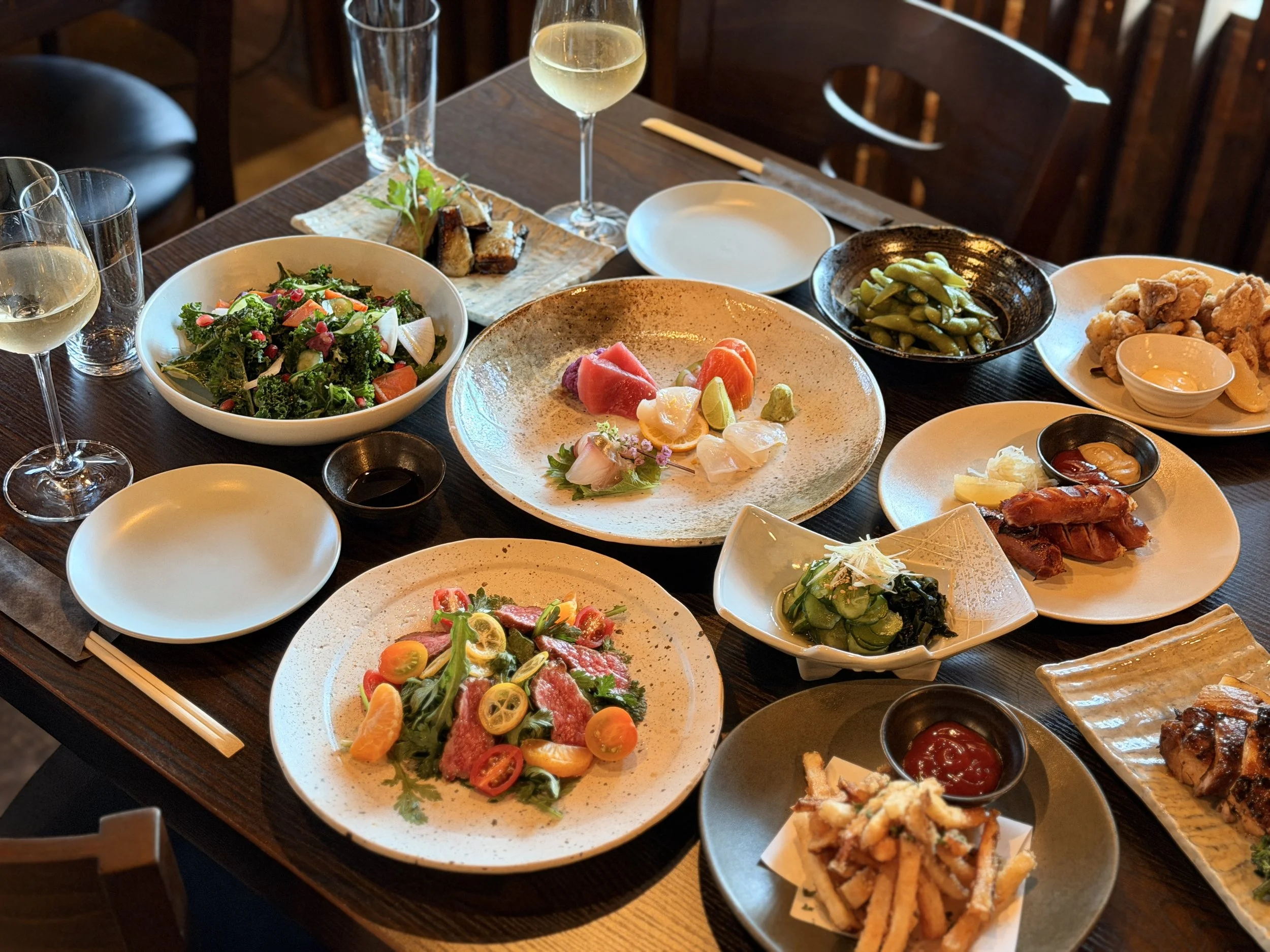 A table filled with various Japanese dishes including sashimi, salad, edamame, fried chicken, and other appetizers, along with glasses of white wine and empty plates.