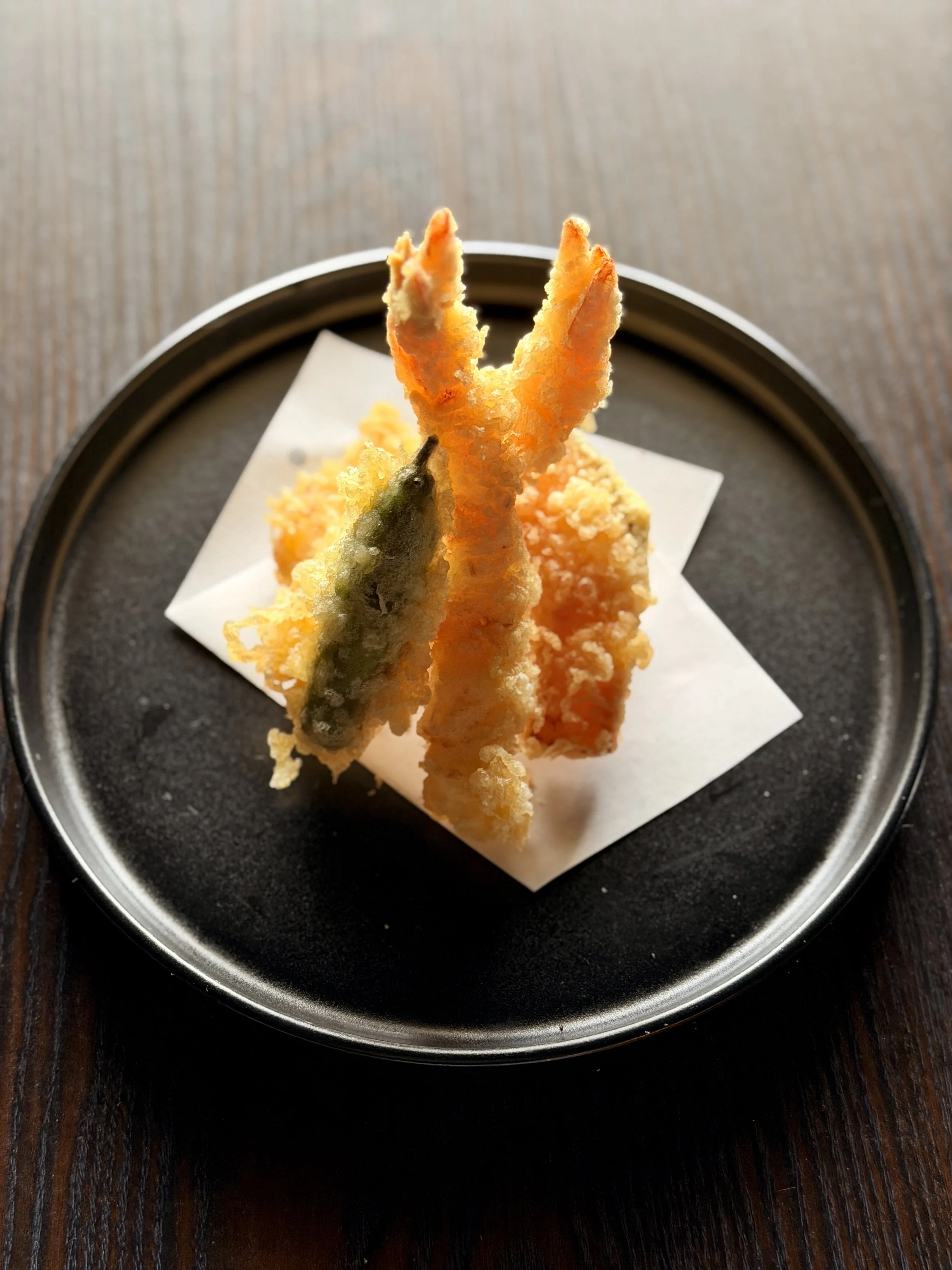 Assorted tempura vegetables and seafood on a black plate with paper, viewed from above.