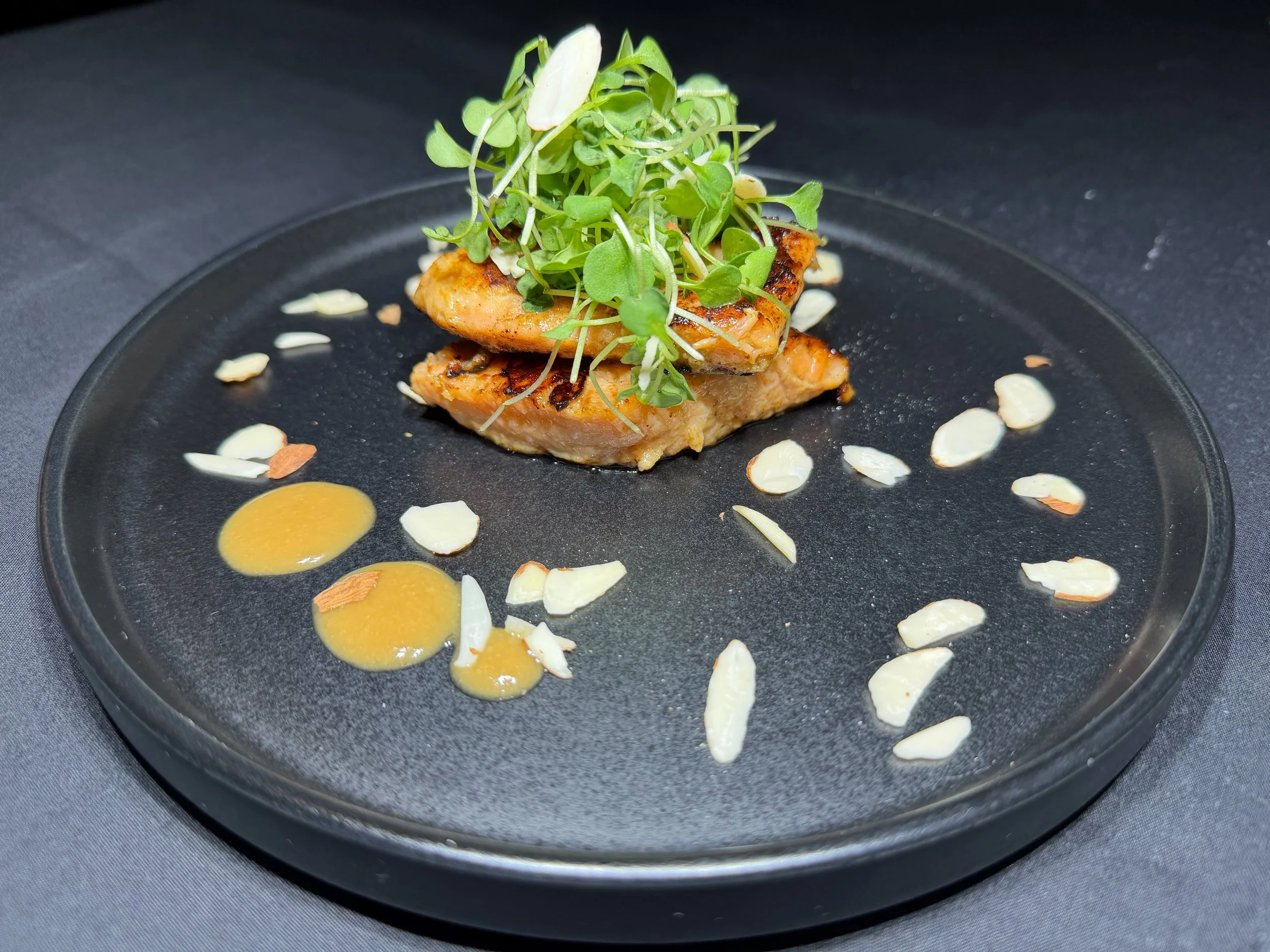 Grilled chicken topped with microgreens, almond slices, and drizzled with sauce on a black plate.