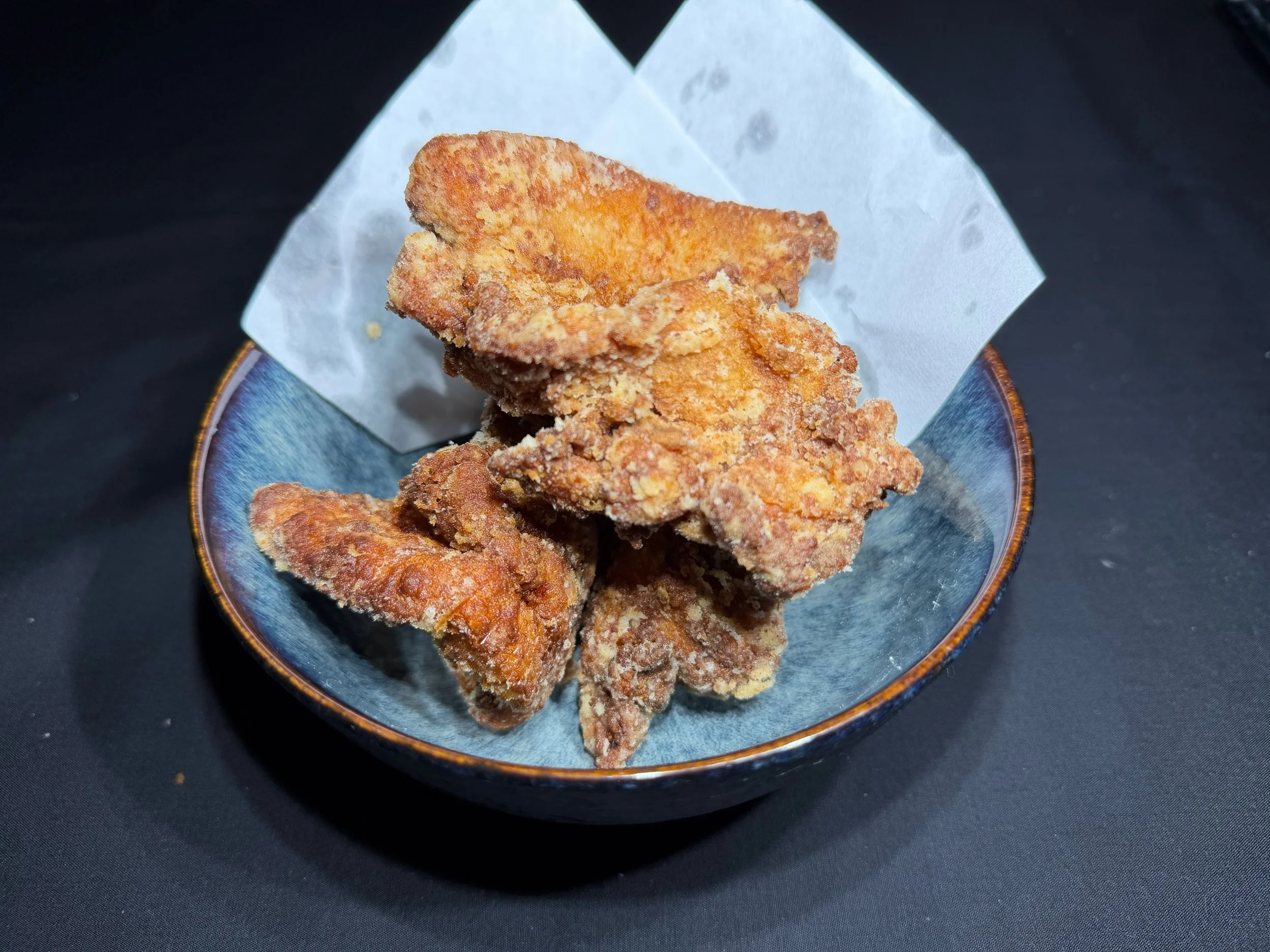 Fried chicken pieces in a small blue bowl with a piece of white paper liner inside, on a black surface.