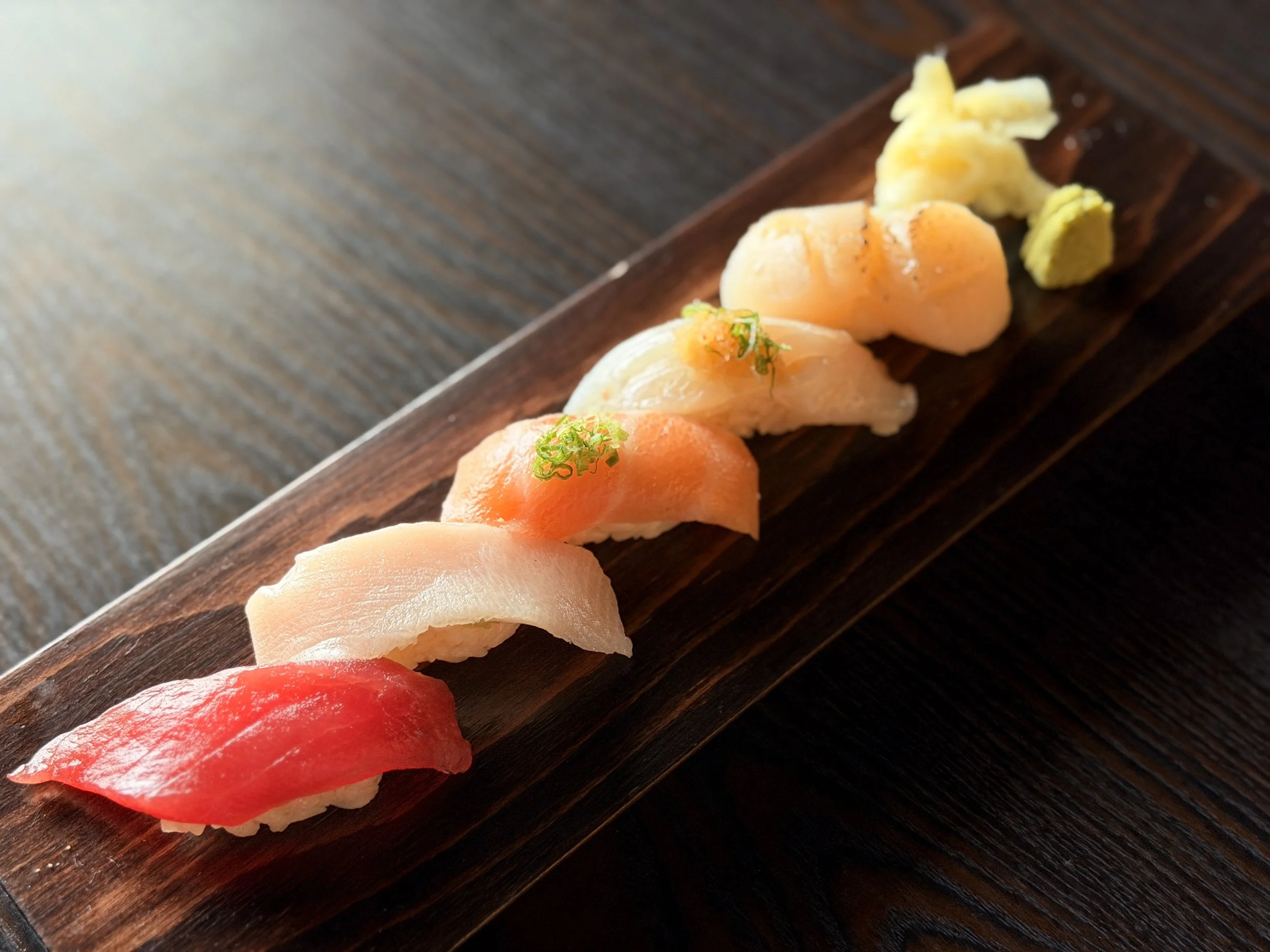 Close-up view of assorted sushi pieces on a dark wooden serving board, including tuna, salmon, yellowtail, and shrimp sushi, with garnishes and wasabi.