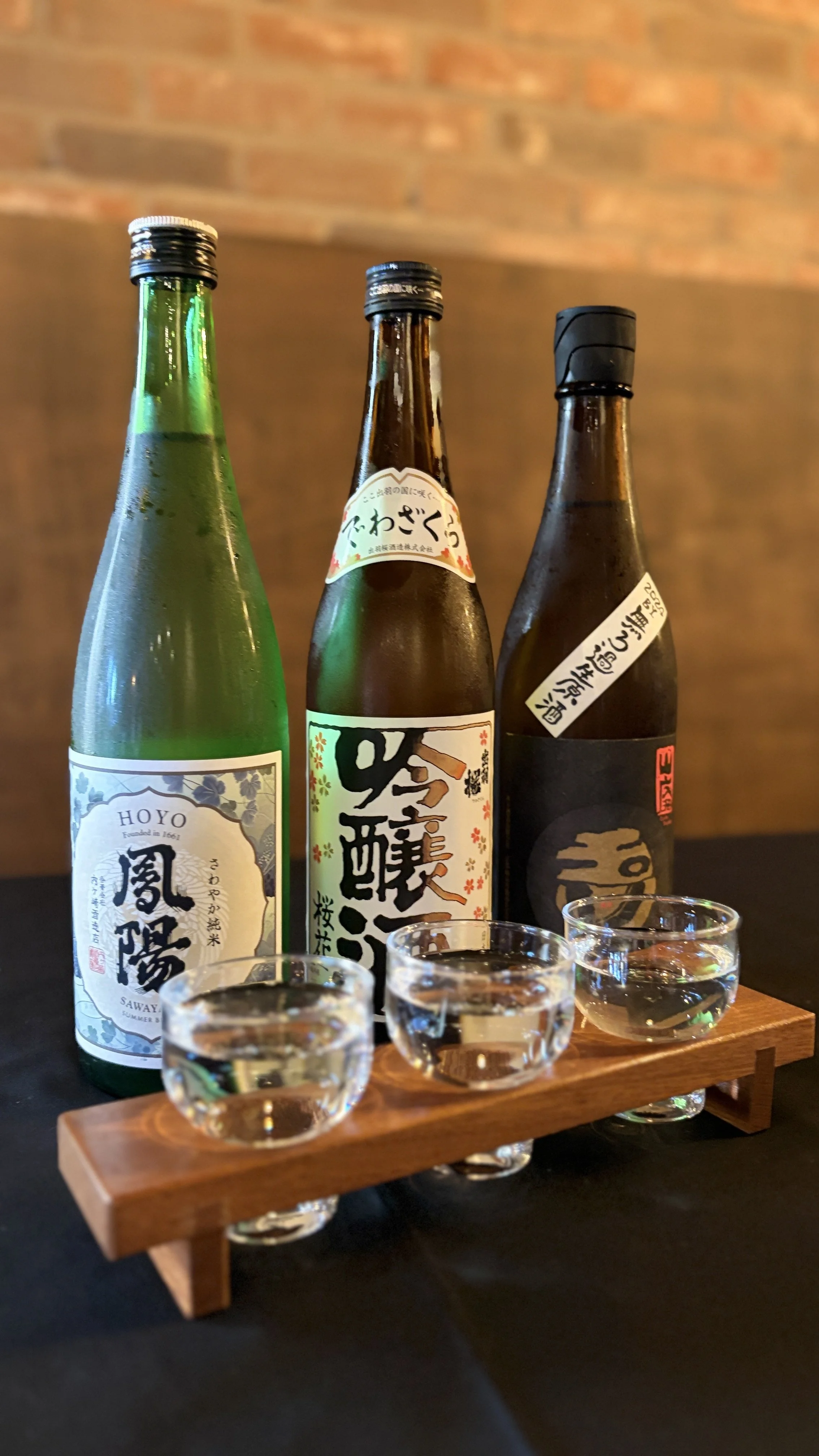 Three bottles of Japanese sake on a wooden stand with four sake cups, set against a brick wall background.