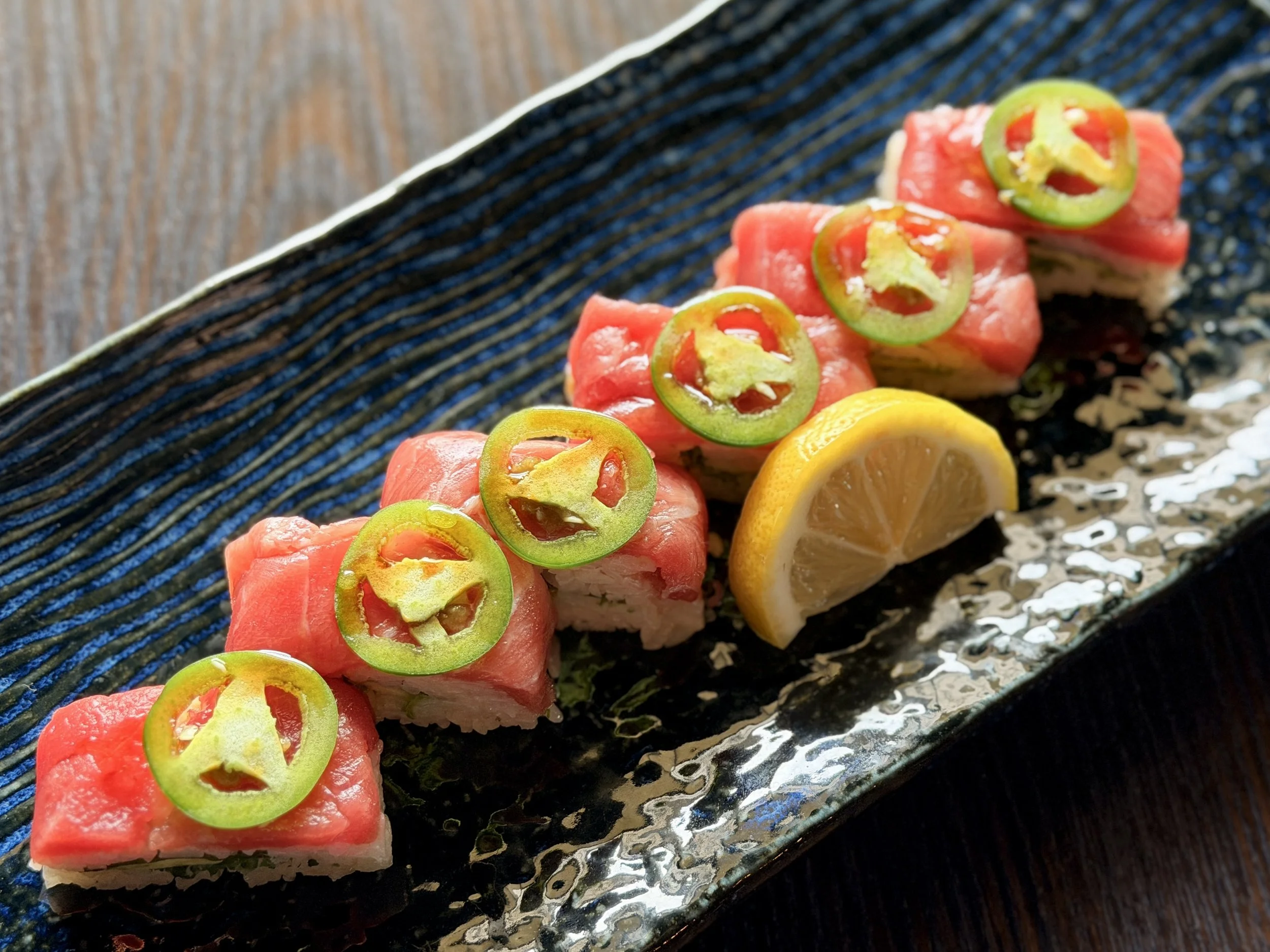 A row of sushi rolls topped with slices of green chili peppers, served on a black textured plate with lemon wedges.