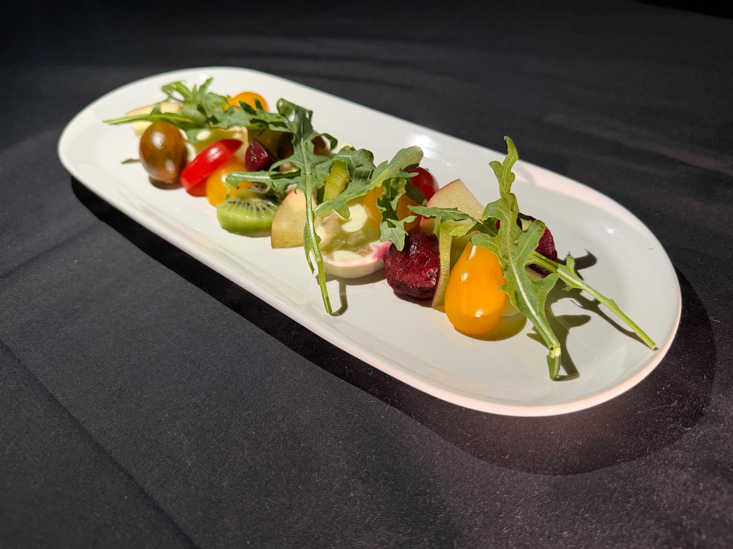 A white oval plate with mixed fresh fruits and greens, including grapes, kiwi, apple slices, cherry tomatoes, and arugula, displayed on a dark surface.