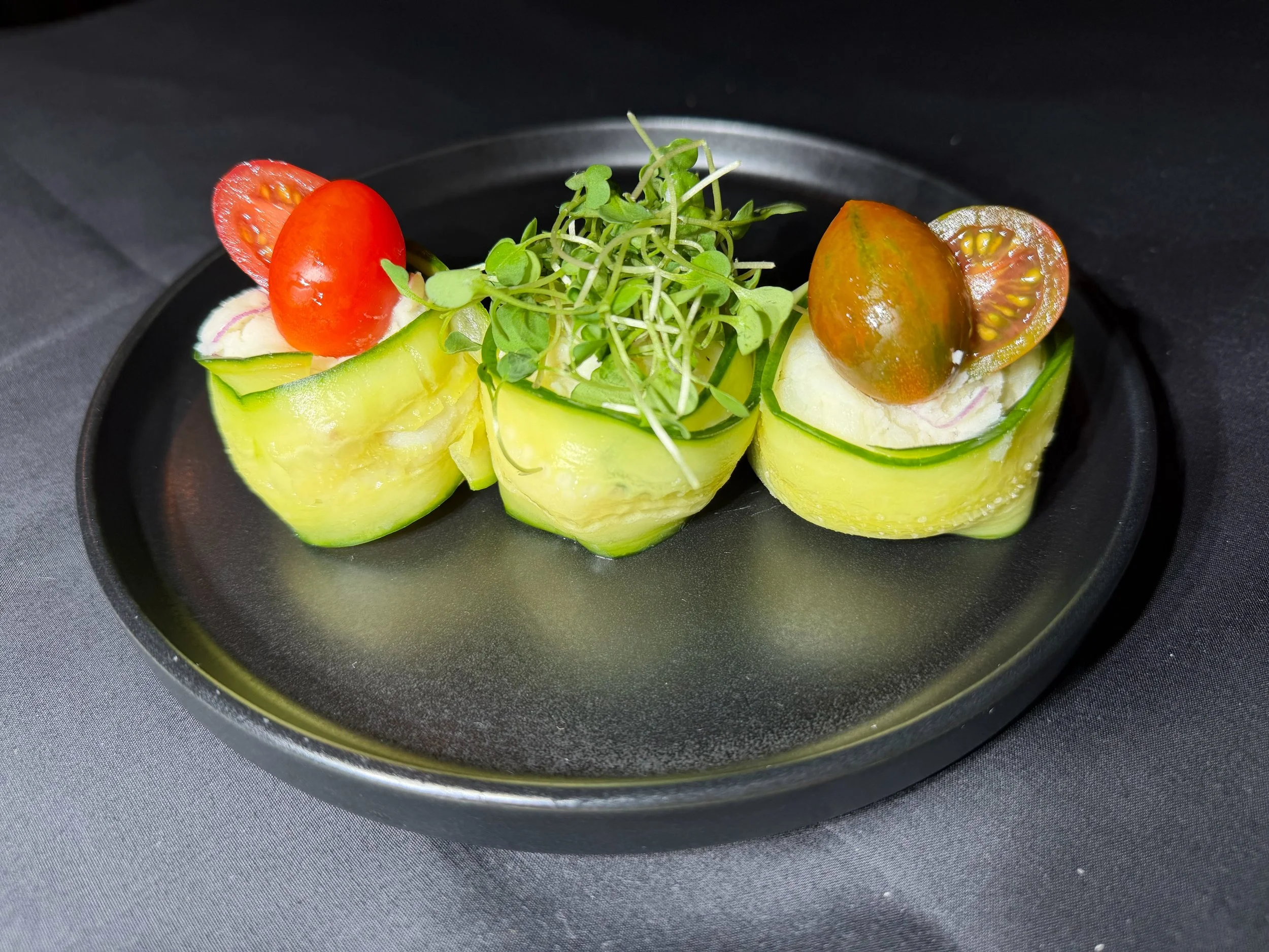 Cucumber cups filled with creamy mixture, topped with cherry tomatoes and microgreens on a black plate.