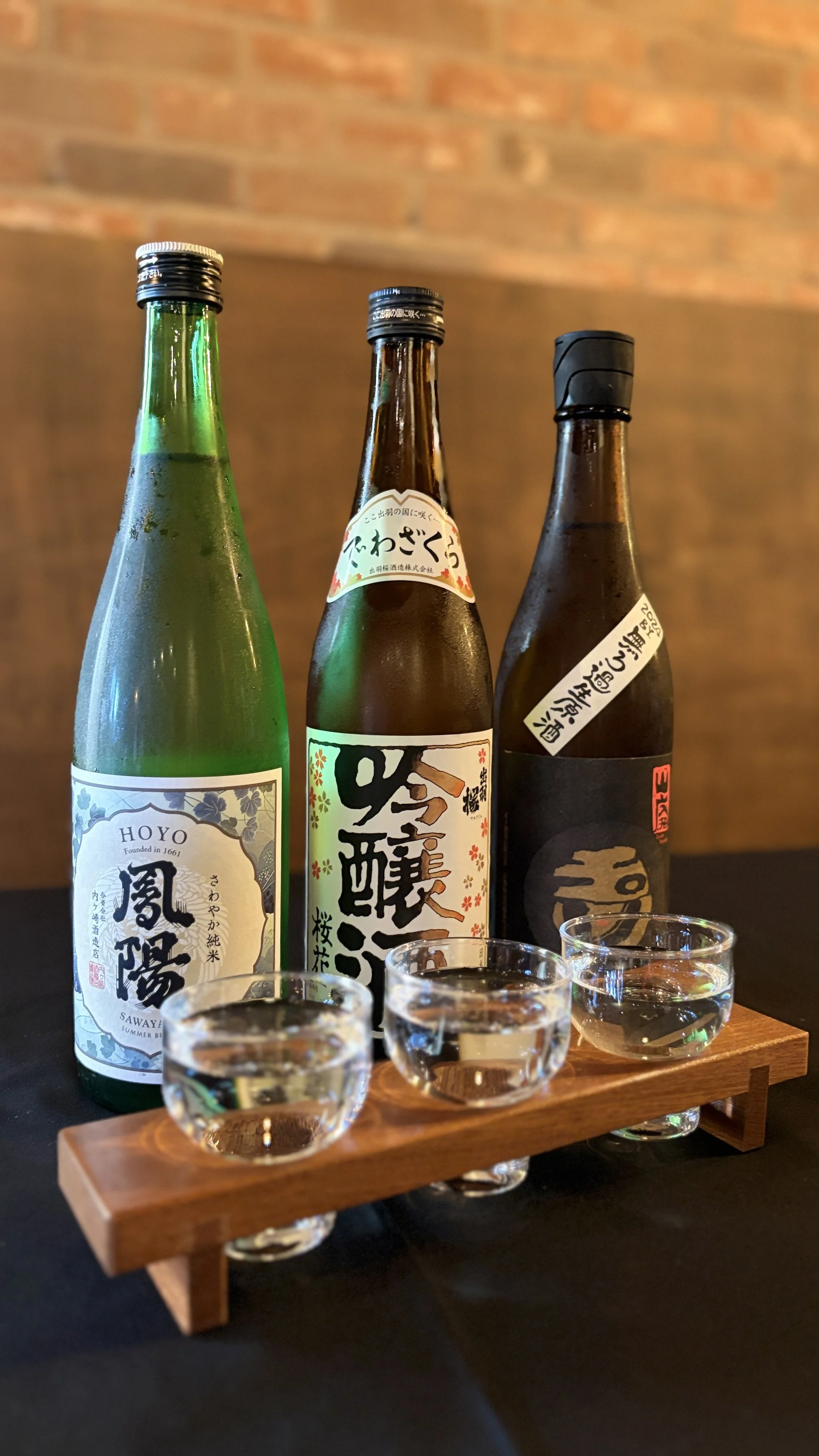 Three bottles of sake on a wooden stand with three small sake glasses, set against a dark surface and brick wall background.