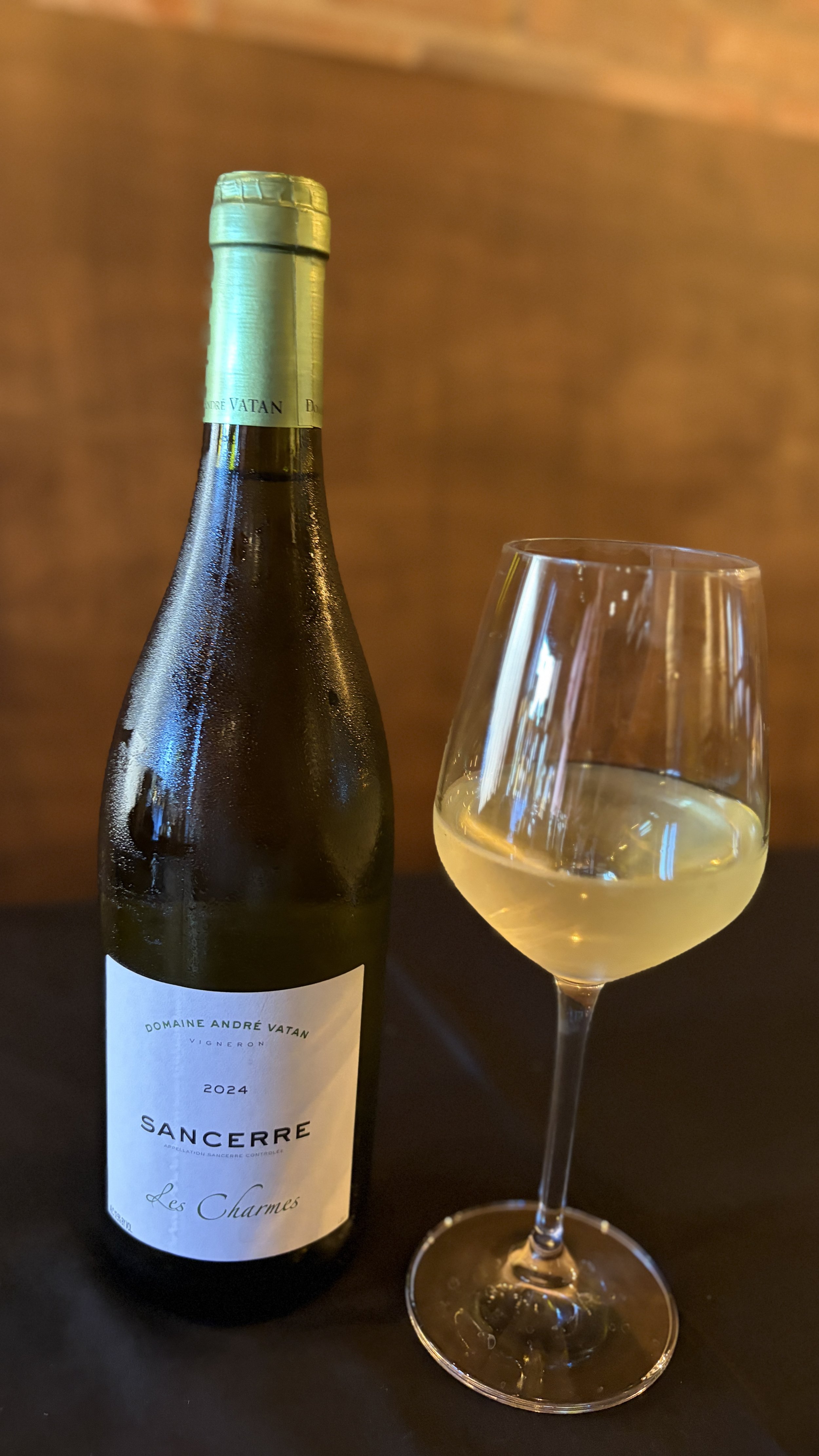 A bottle of Sancerre Les Charmes white wine and a filled wine glass on a dark surface against a warm brown background.