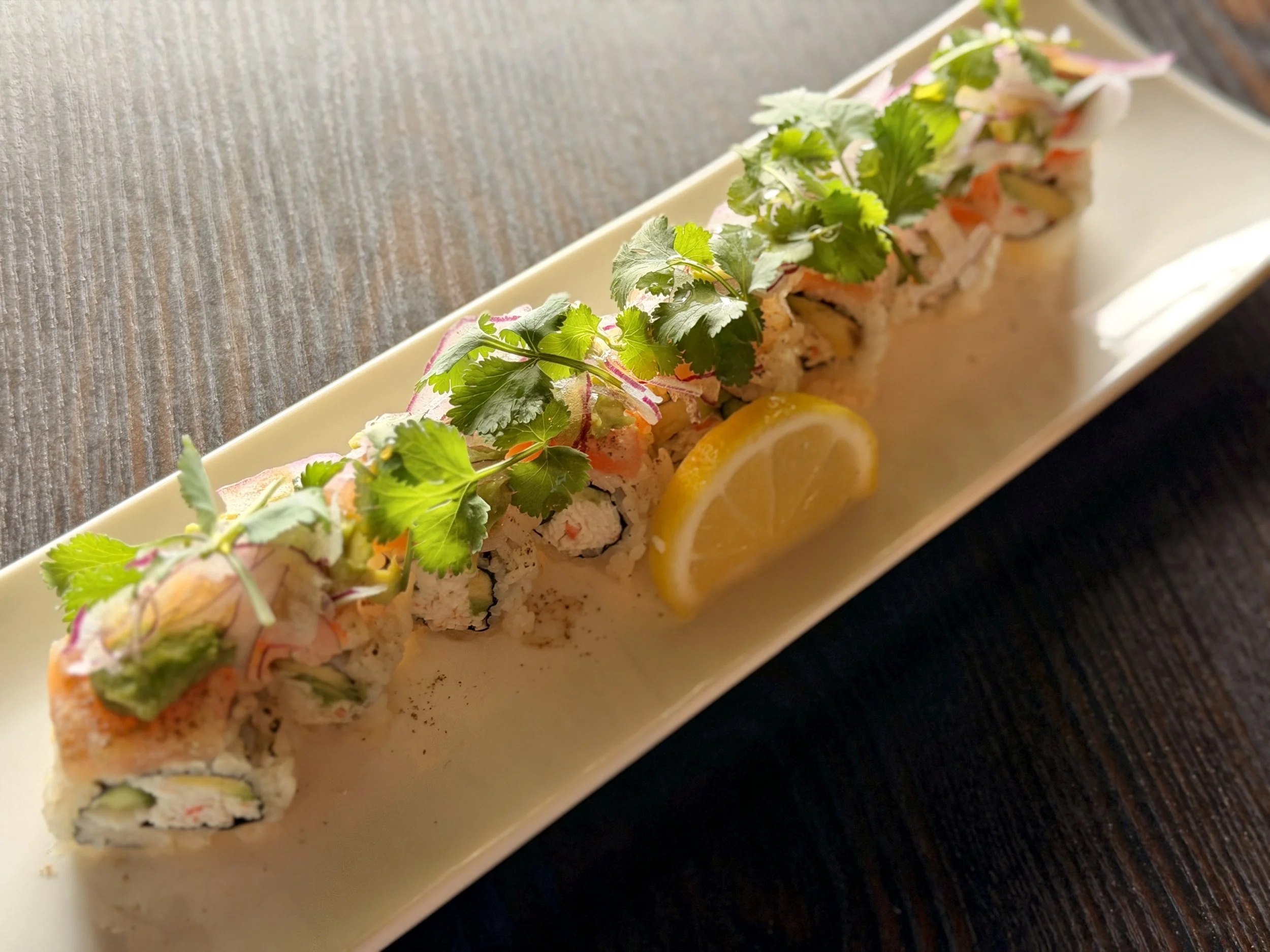 A long rectangular plate of sushi with various types of rolls topped with herbs and slices of lemon, resting on a dark wooden surface.