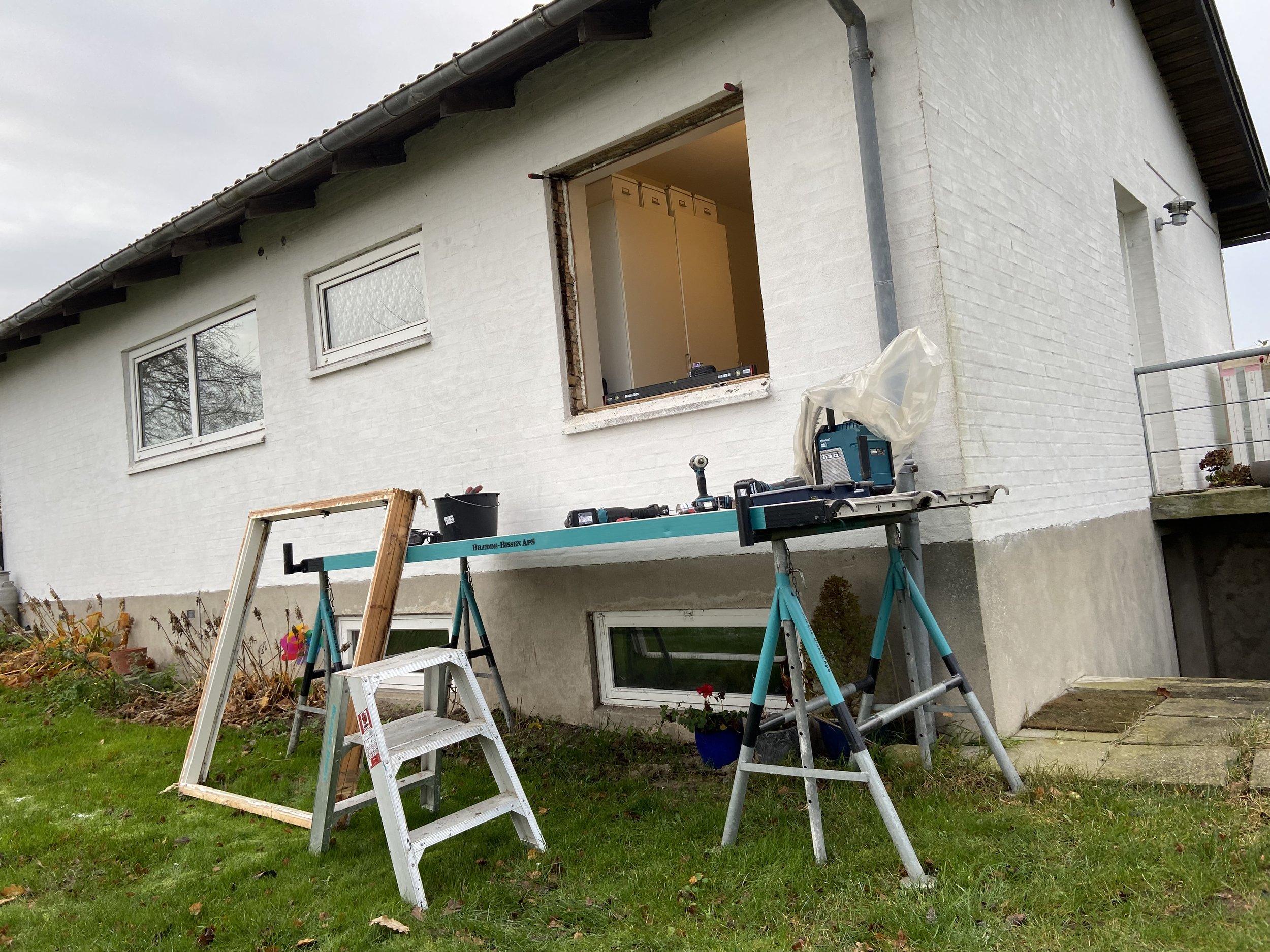 Udendørs billede af en hvid husfacade under vinduesudskiftning, med værktøj og stillads foran, herunder en trappestige.