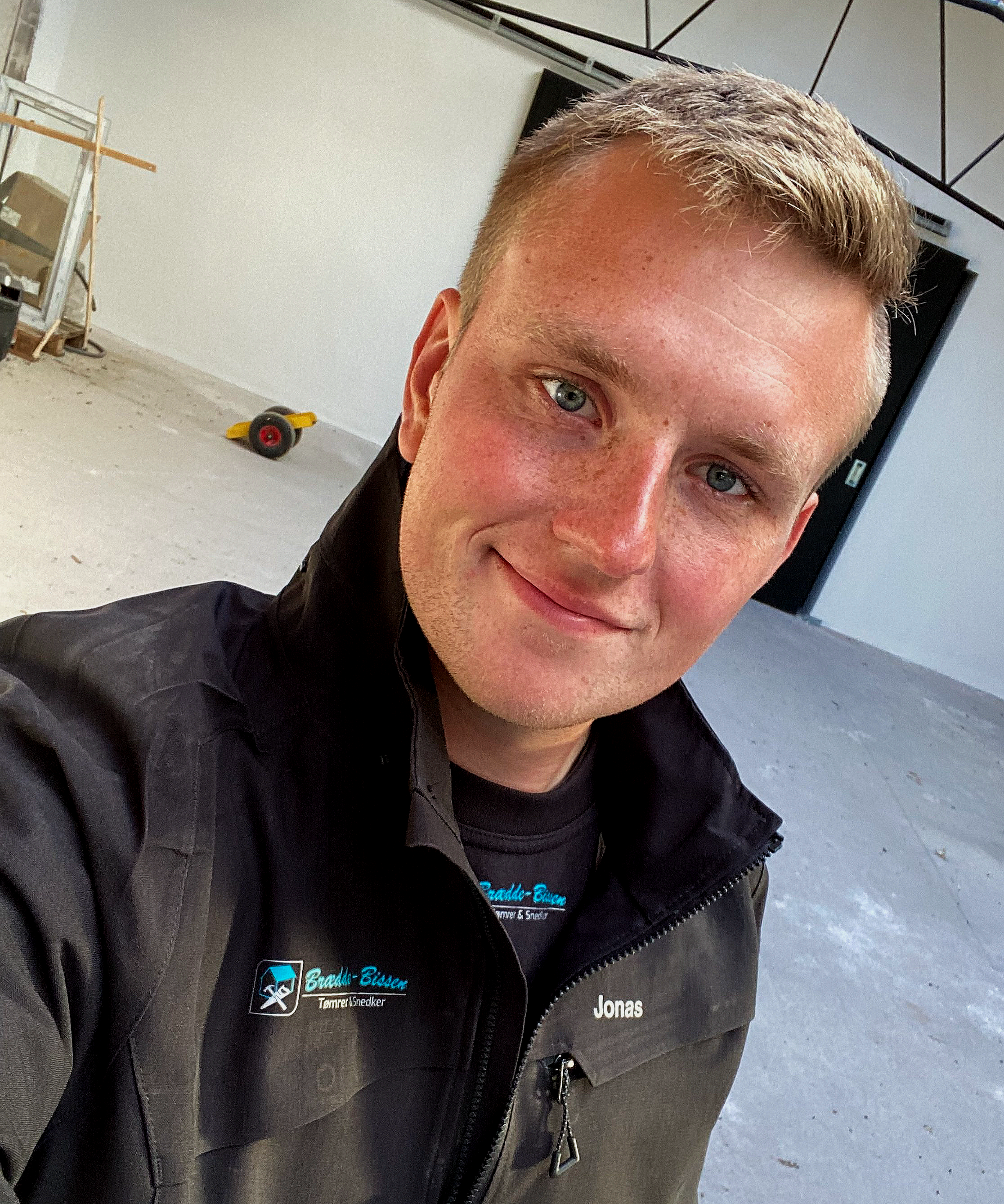 En ung mand med blondt hår og blå øjne, smilende, iført sort jakke med navn 'Jonas' og logo for 'Breddi-Bisser'. Han står i et tomt, indendørs rum under renovering med en gul værktøjsvogn og byggematerialer i baggrunden.