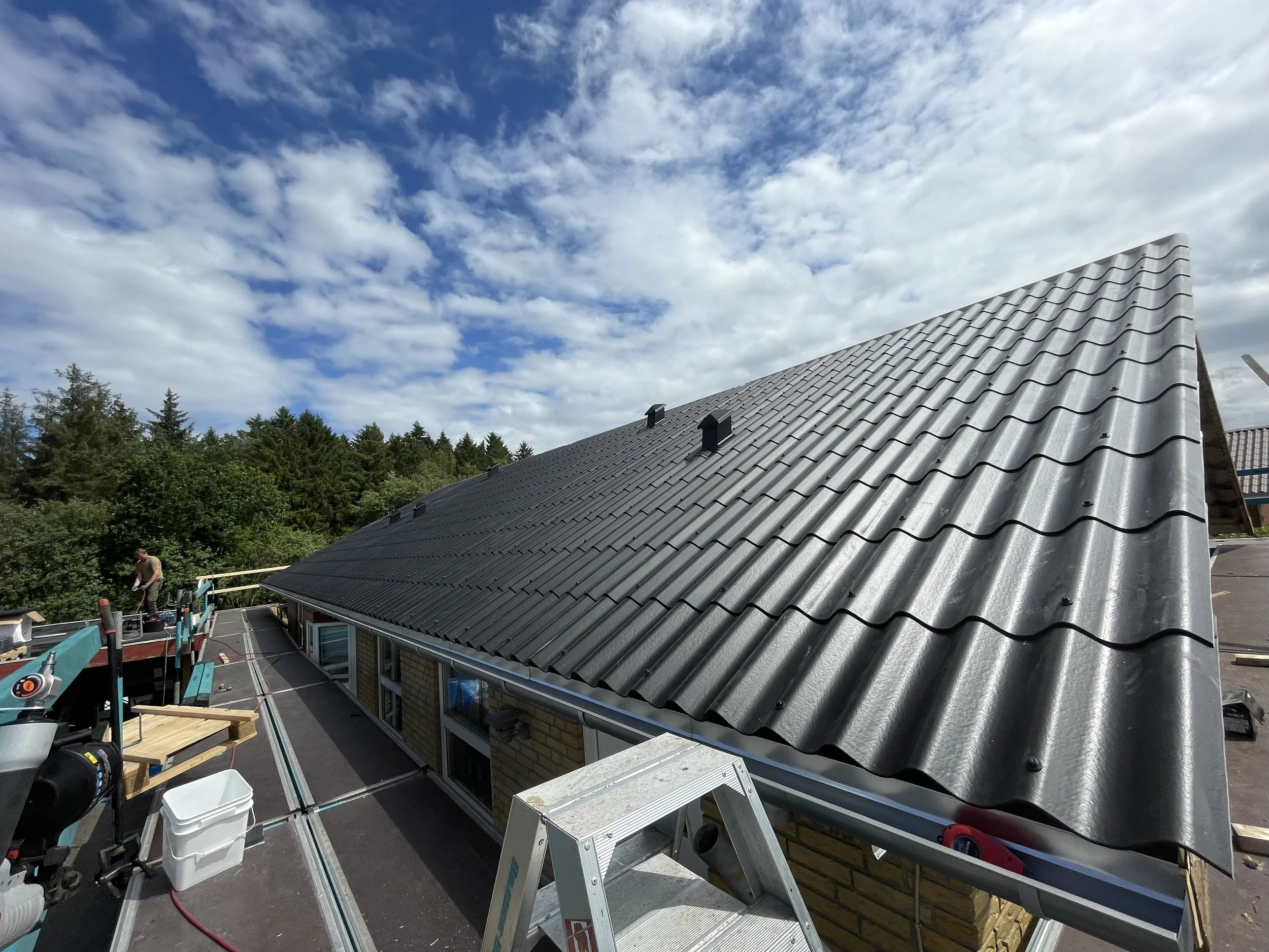 Et tag under renovering med sorte tagskifer, arbejdsudstyr og en person, der arbejder på taget, med en skov i baggrunden og en skyet himmel.