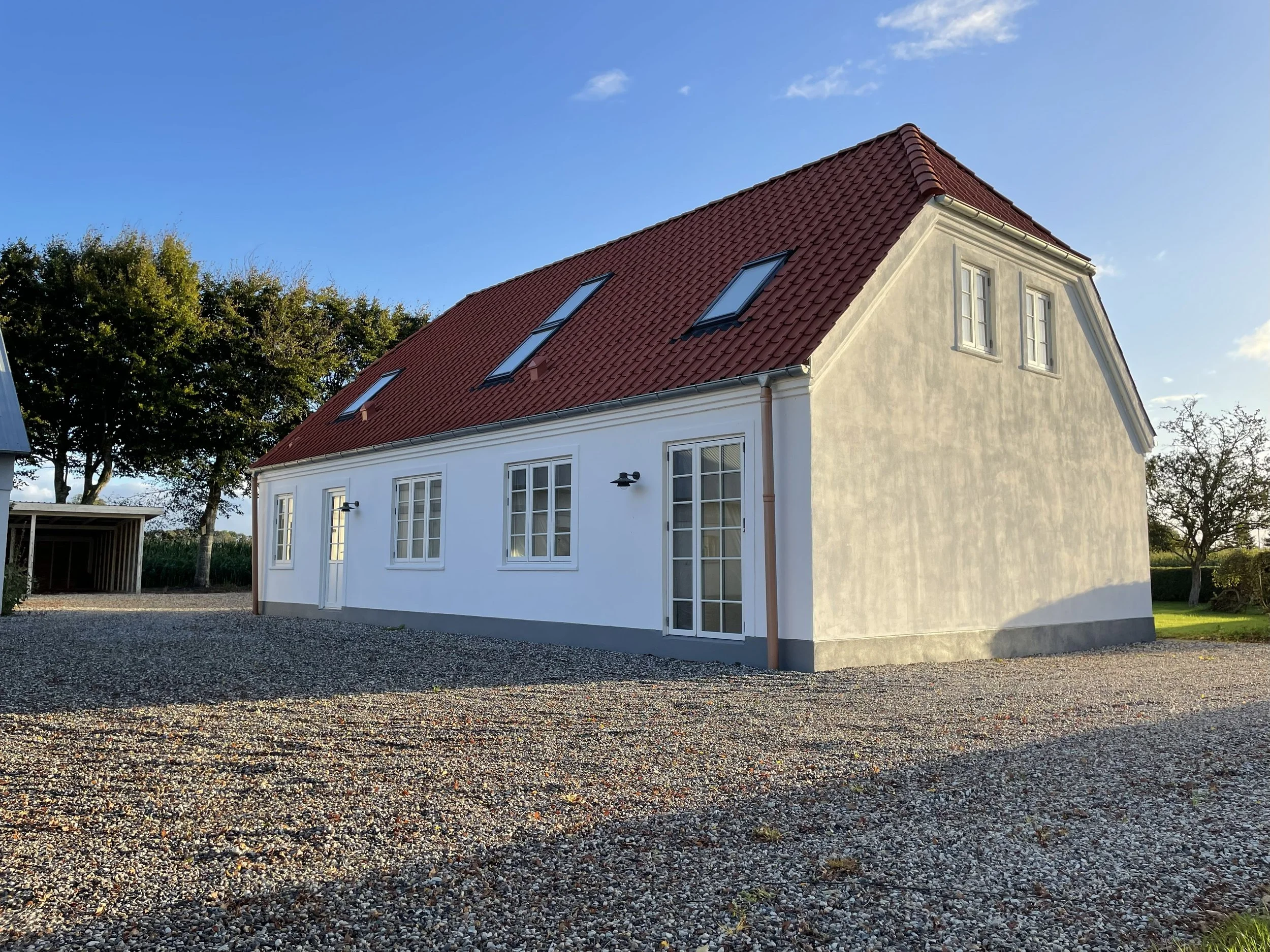 Et hvidt hus med rød tagstens-tag, flere vinduer og to udendørs væglamper, omgivet af en grusbelagt indkørsel og et haveområde med træer under en blå himmel.