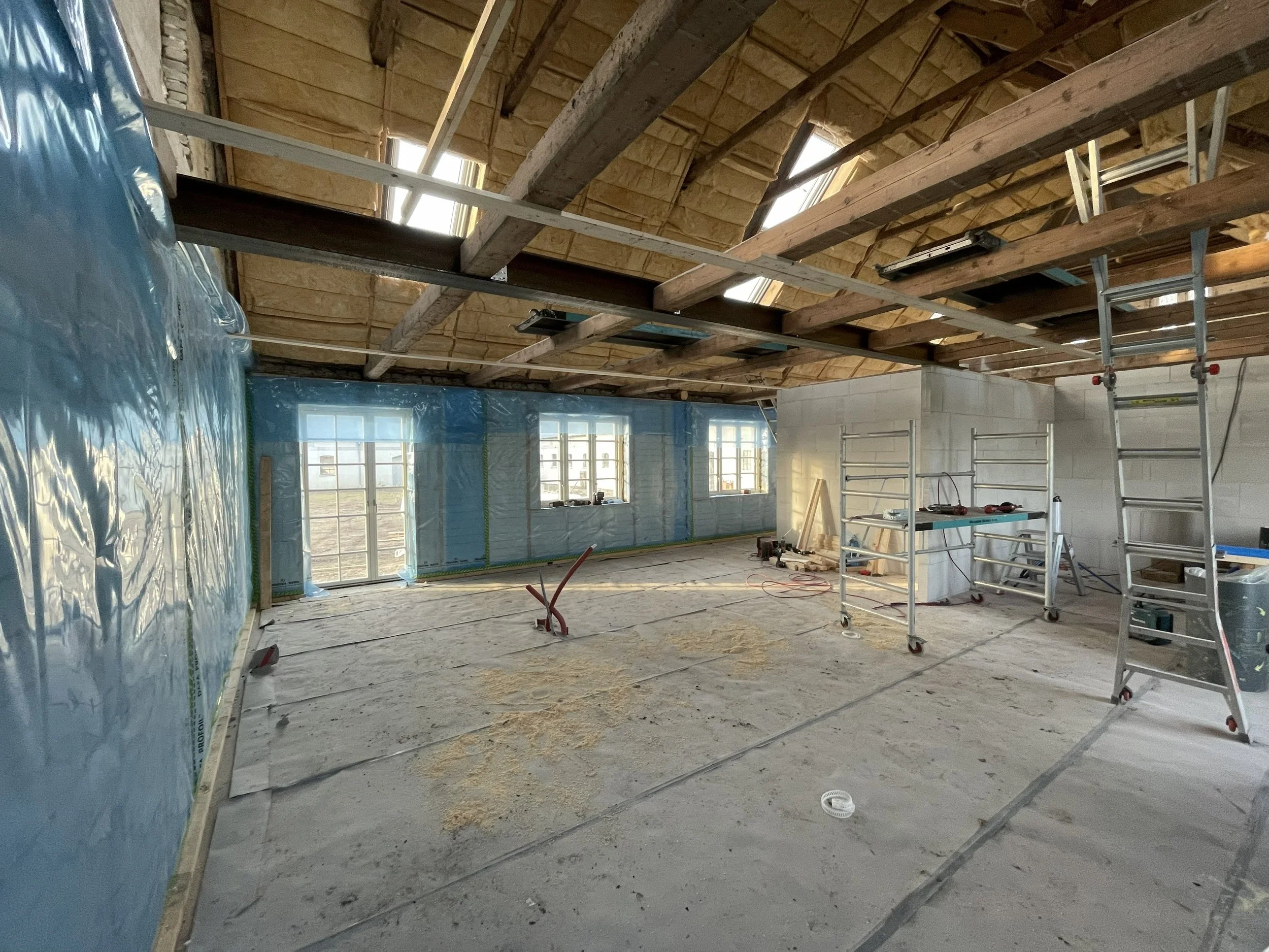 Indendørs byggeplads med poleret betongulv, vægge dækket med blå dampspærreplastik, flere vinduer, loft med synlige trætømmer, og skinner samt stillads til arbejde. Der er materialer og værktøj spredt rundt.