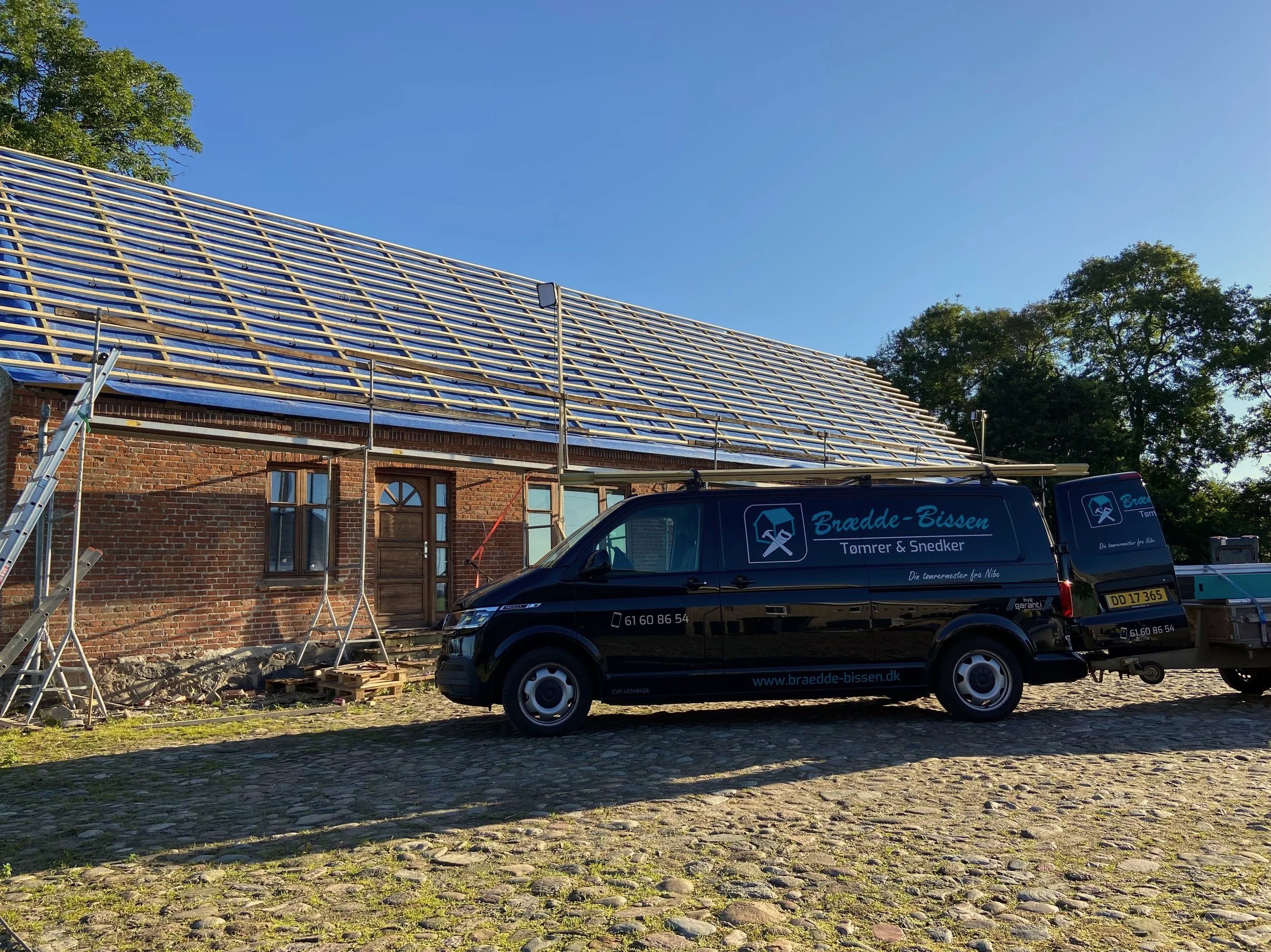 En sort firmabil med teksten 'Brædde-Bissen Tømrer & Snedker' parkeret foran en gammel murstensbygning under opførelse med nyt tag. Der er udstyr og værktøj til tagarbejde.