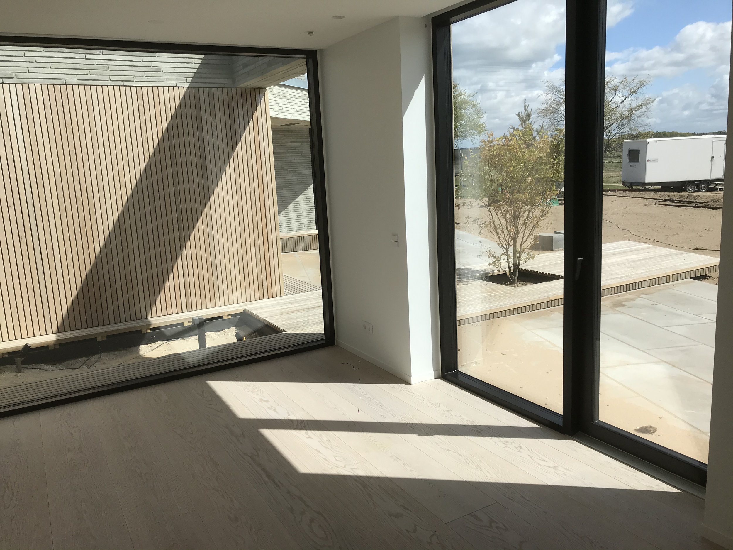 Indendørs rum med store gulv-til-loft vinduer, der vender ud mod en træterrasse udført i egetræ og en lille træbusk udenfor.