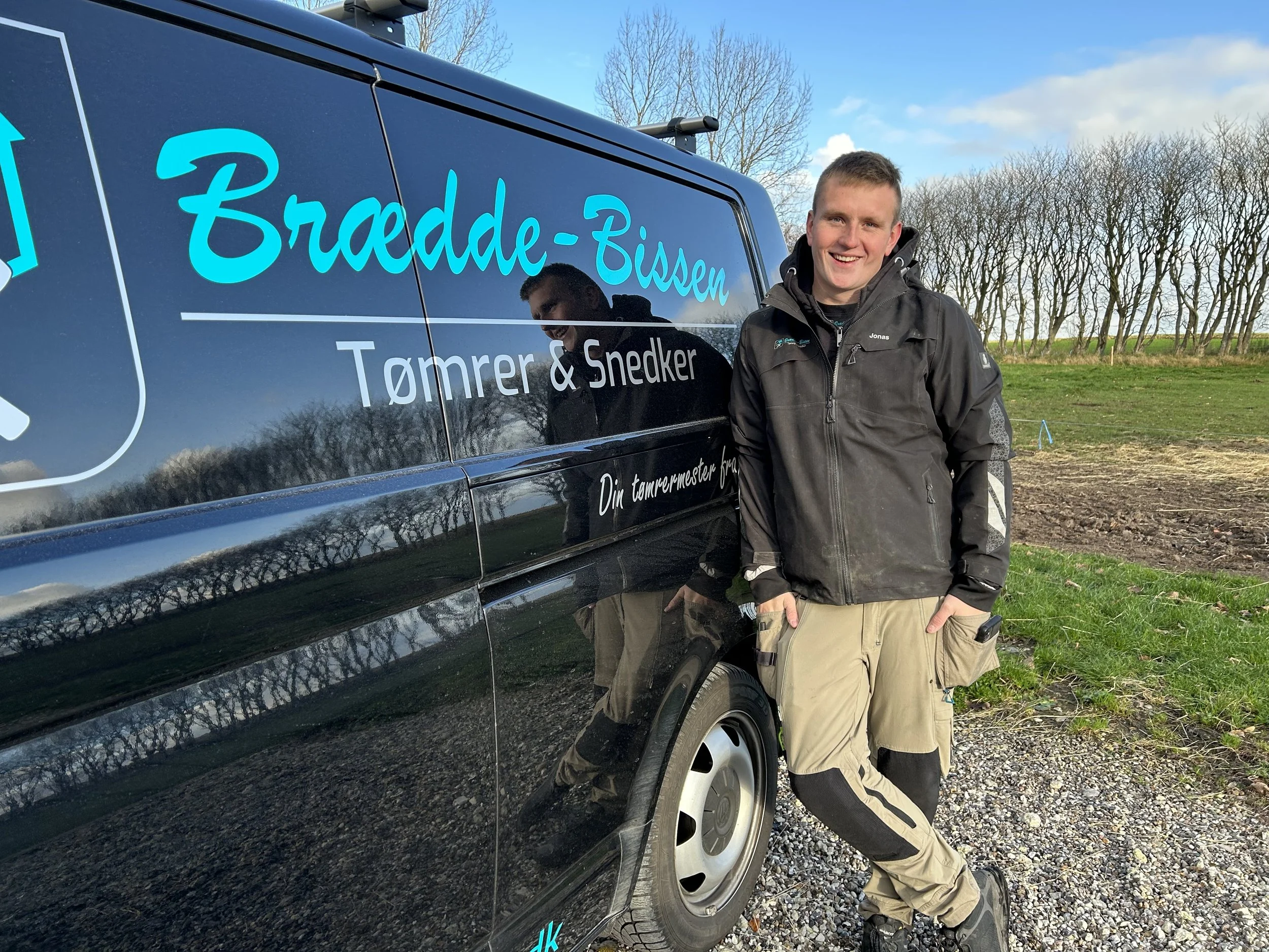 Ung mand står ved siden af en sort firmabil med teksten "Bredde-Bissen Tømrer & Snedker" og en undertitel, der siger "Din tømrer mester fra Djørup" på en åbengræsmark med træer i baggrunden, under en blå himmel med nogle skyer.