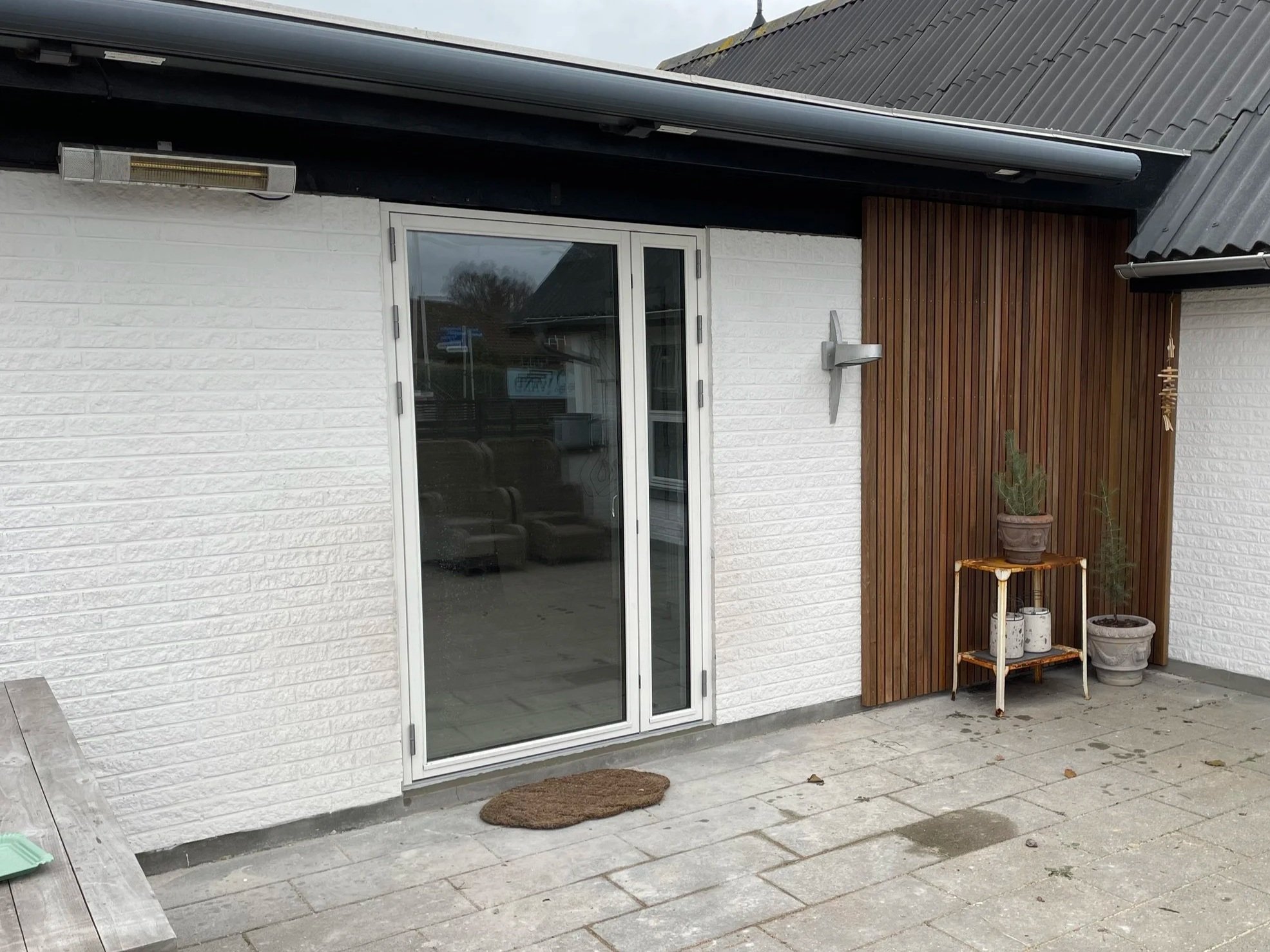 Udsigt af hus, indeholder en glasdør med havedør, hvide murstensvægge, træpanel på væg samt små havepotter med planter.