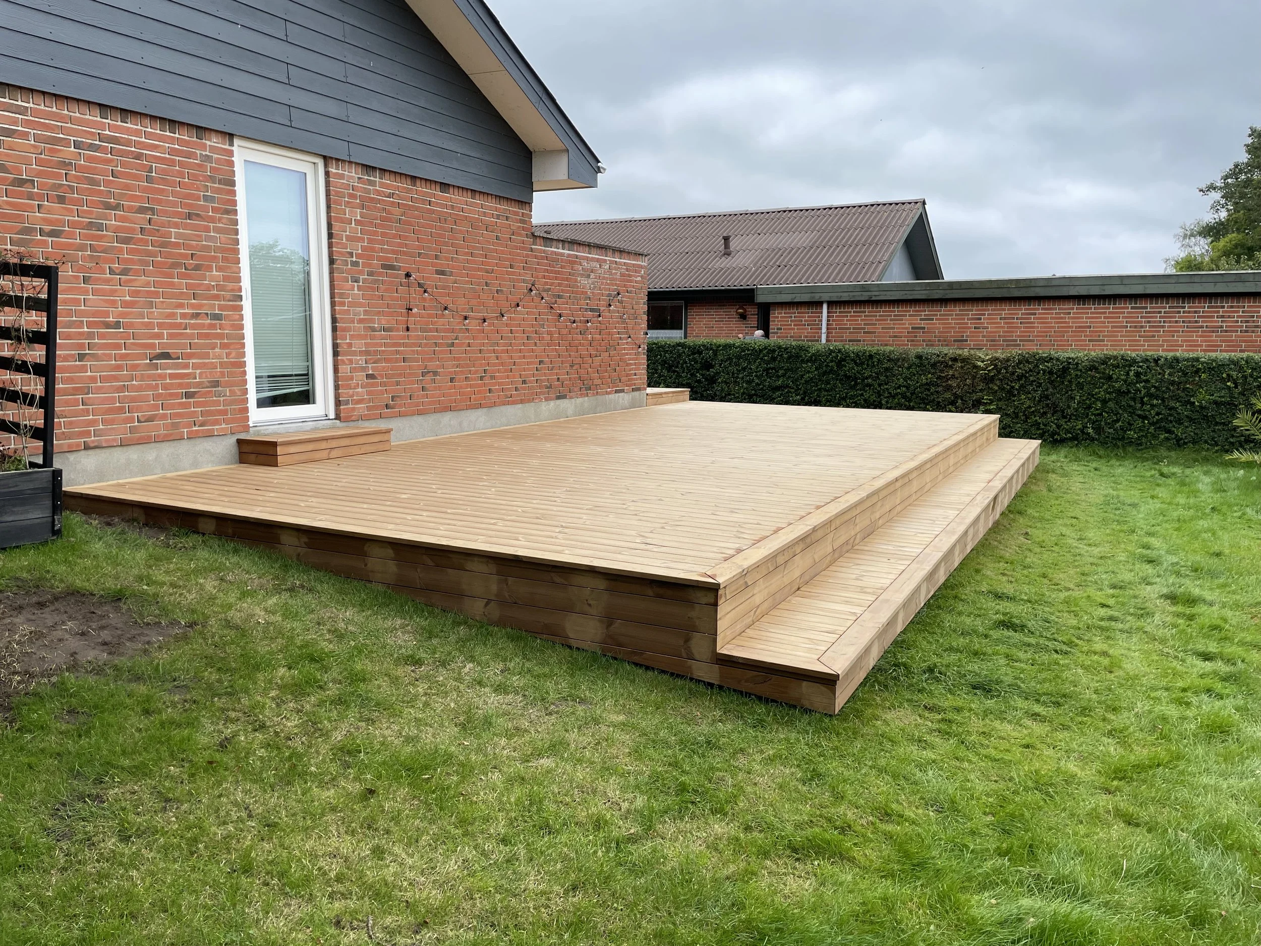 En træterrasse udenfor en murstenshus med baghave, græsplæne og hængende lysstrenge.