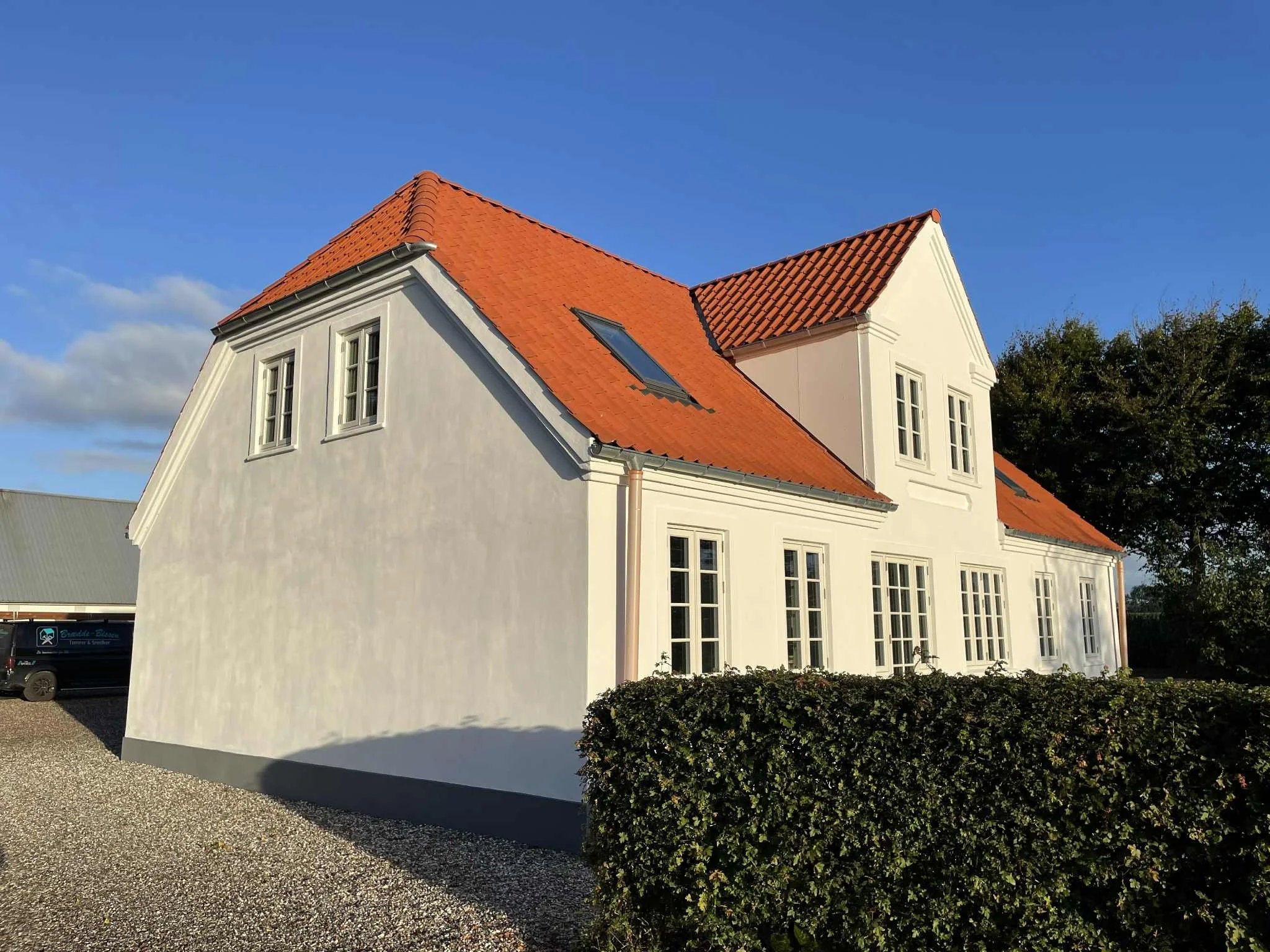 Hvidt hus med rødt tegltag og mange vinduer, omgivet af grøn busk og blå himmel.