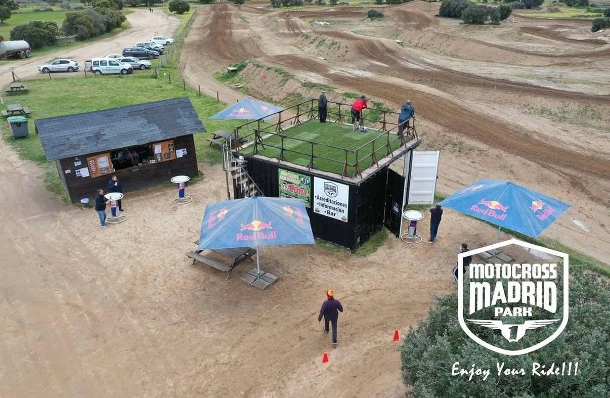Área de parqueo y zona de espectadores en una pista de motocross en Madrid, con personas observando y tomando fotos, toldos con logotipo de Red Bull, estructura con césped artificial y vista de la pista de tierra para carreras.