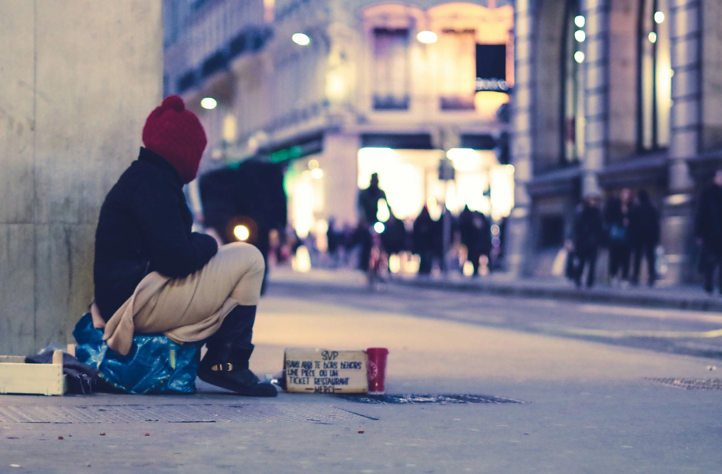 A person wearing a red knit hat, black jacket, and beige pants sitting on the sidewalk by a wall, with a small sign and cup nearby, in an urban area during evening or nighttime with blurred city lights and pedestrians in background.