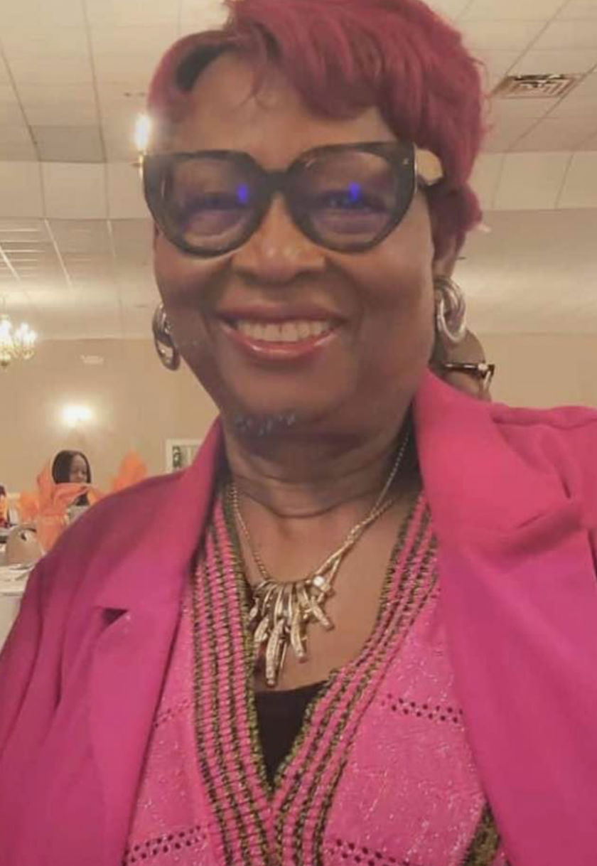 A woman smiling at a social event, wearing large sunglasses, a pink blazer, and a pink top with intricate details. She has short, red hair, hoop earrings, and a layered necklace.