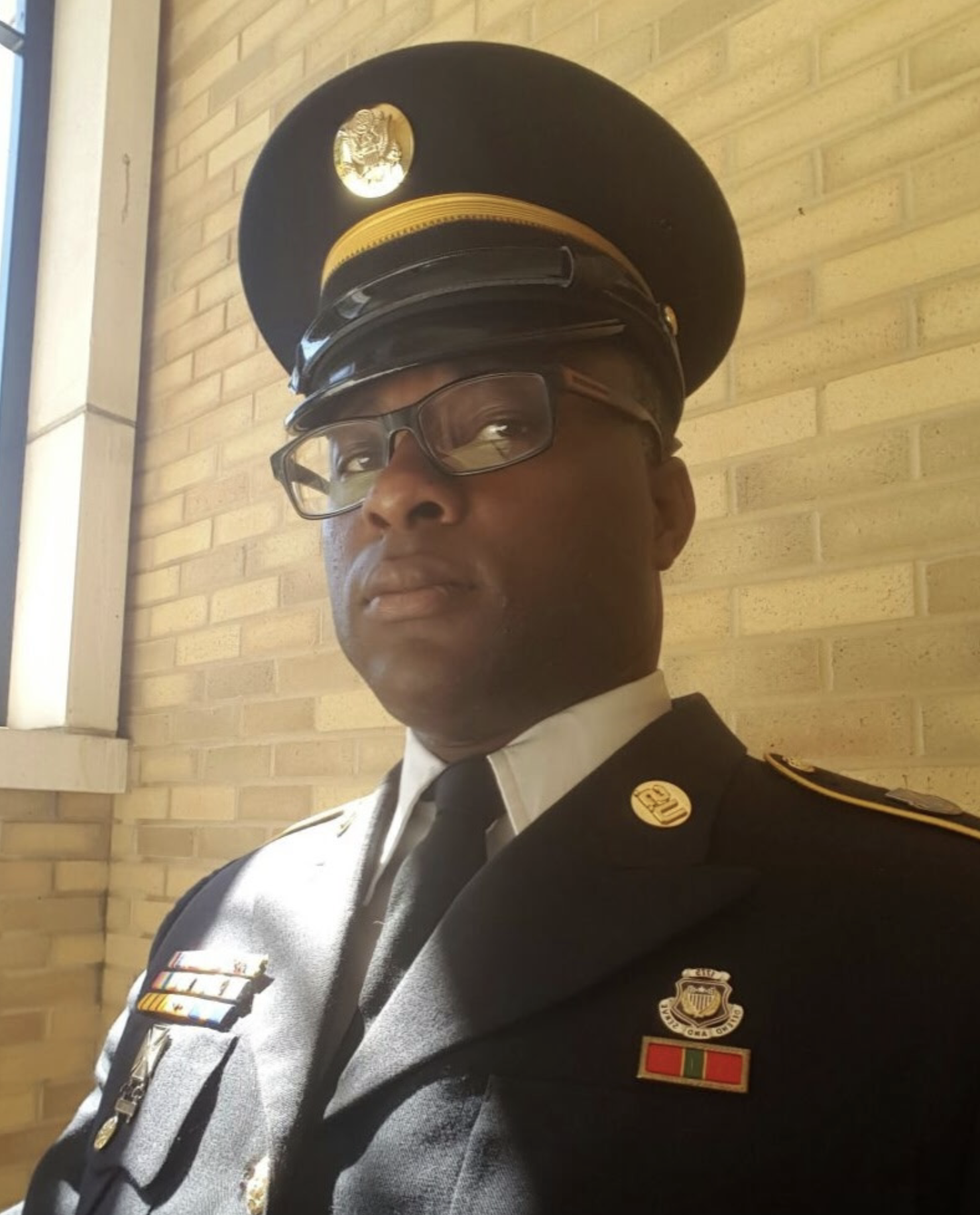 A man in a U.S. military uniform with medals and a hat, standing indoors against a brick wall.