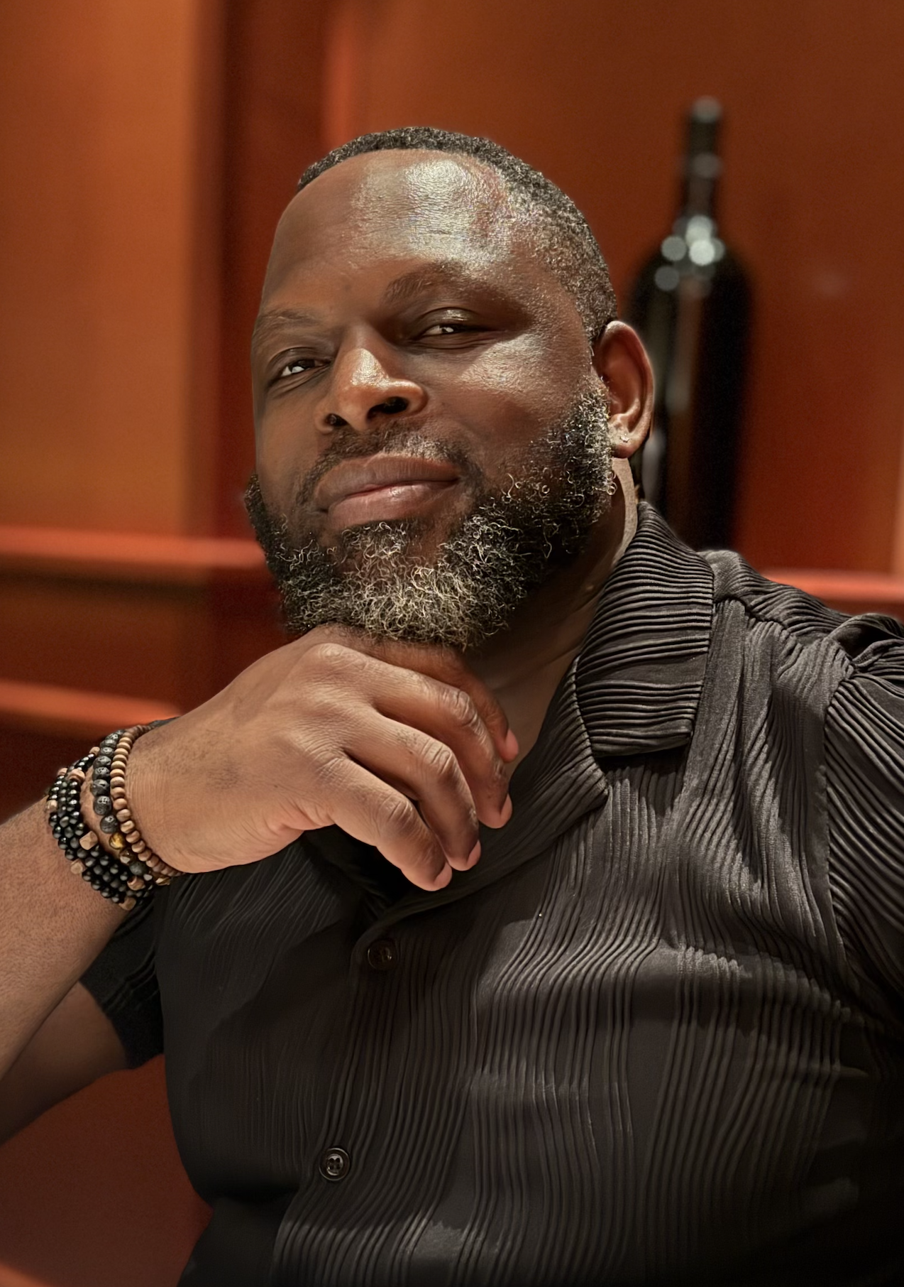A man with a beard and short hair, wearing a black, ribbed, button-up shirt, sitting indoors with a relaxed pose and a thoughtful expression, with a blurred wooden background and a dark, abstract sculpture in the background.