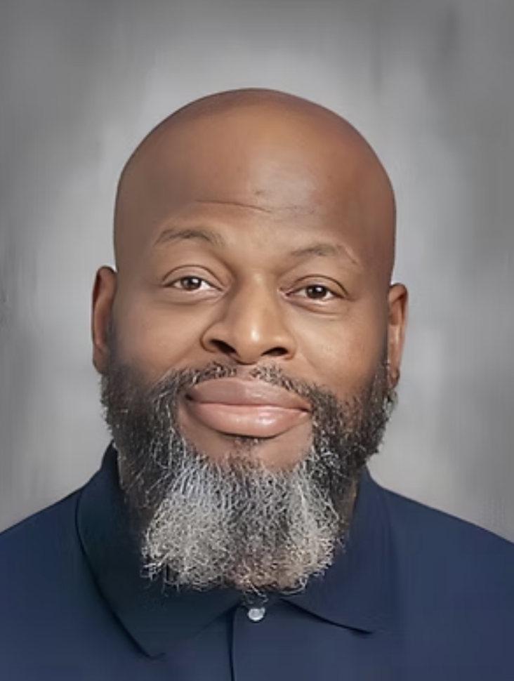 Close-up portrait of a bald man with a gray beard, wearing a dark collared shirt, smiling softly, against a neutral gray background.