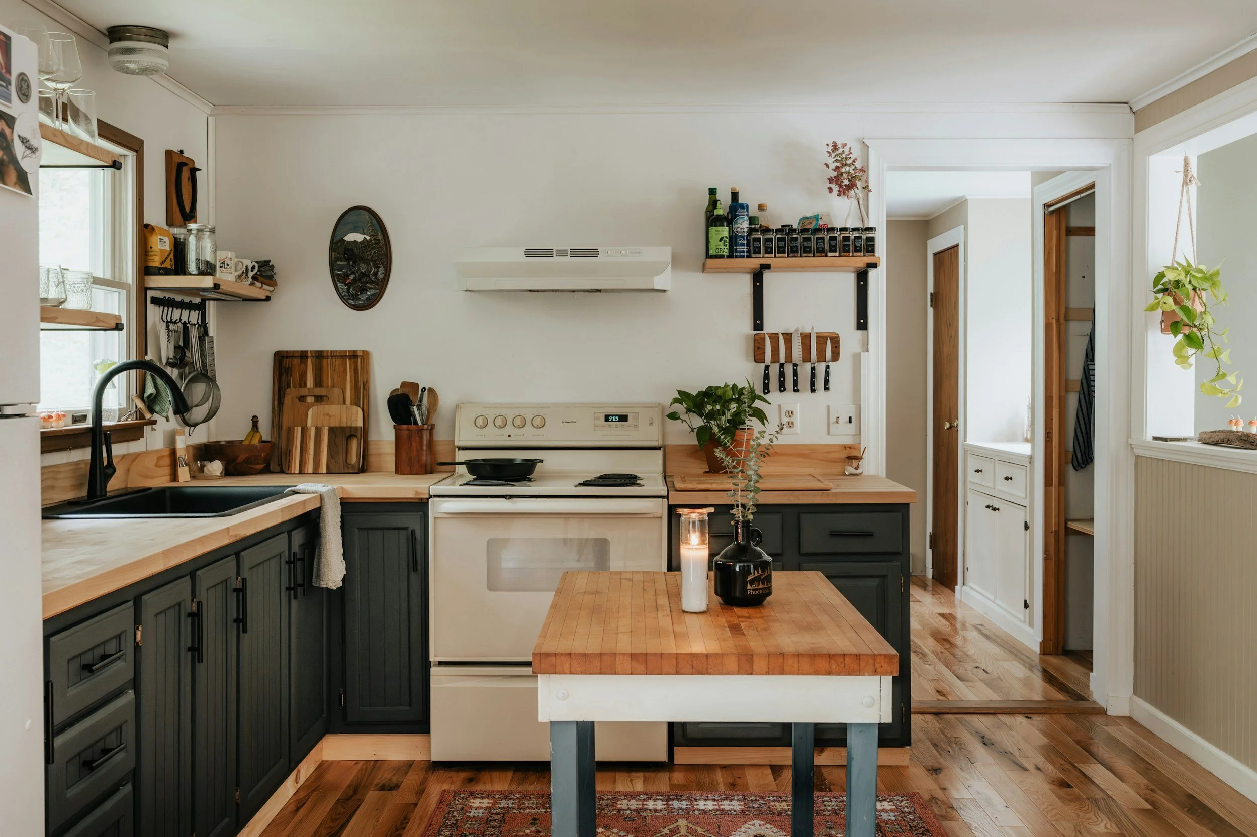 A kitchen with white walls, wooden and black cabinetry, a white stove, cutting boards, and plants on the counters and shelves.
