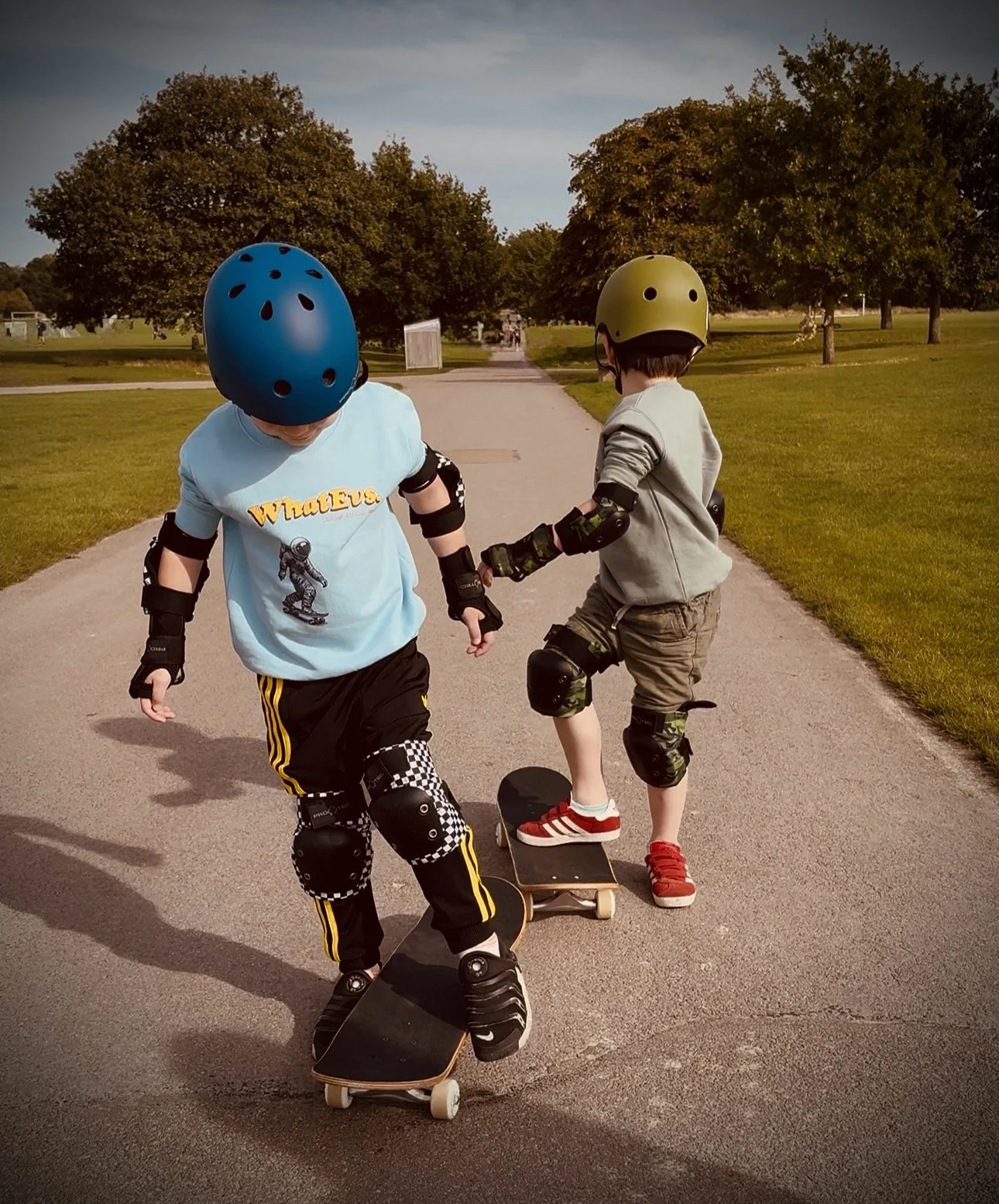 Two children riding skateboards in a park with green grass and trees, wearing helmets and knee pads, holding hands.