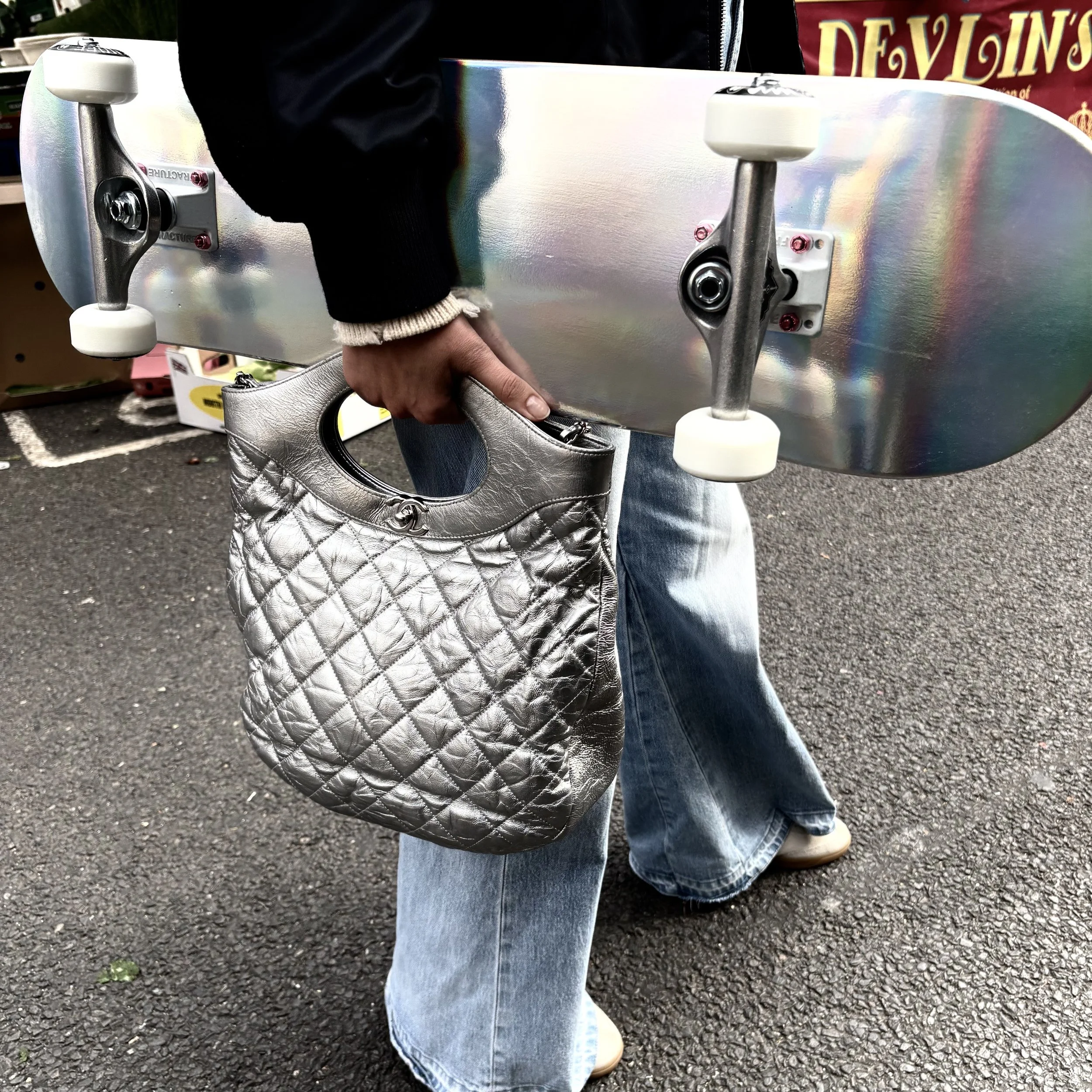 Person holding a silver metallic quilted handbag and skateboard, wearing jeans, standing on a paved surface outdoors.