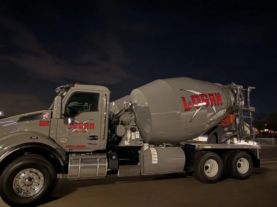 A cement mixer truck with the word LOGAN on the drum parked on a dark street at night.
