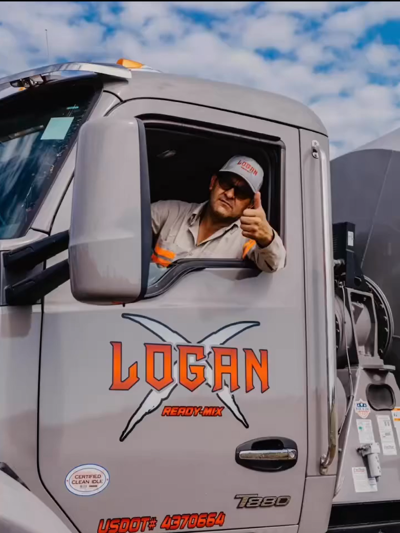 A man wearing sunglasses, a gray cap with red text, and a light-colored shirt with reflective orange accents is sitting in the driver's seat of a large gray truck. The truck has the word "LOGAN" in large orange letters with a black and white X behind