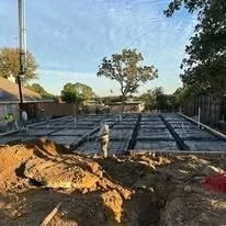 Construction site with foundation pouring in progress, surrounded by dirt and construction materials, with trees and a building in the background.