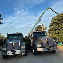 Two large black dump trucks are parked outdoors with a clear blue sky and green trees in the background.