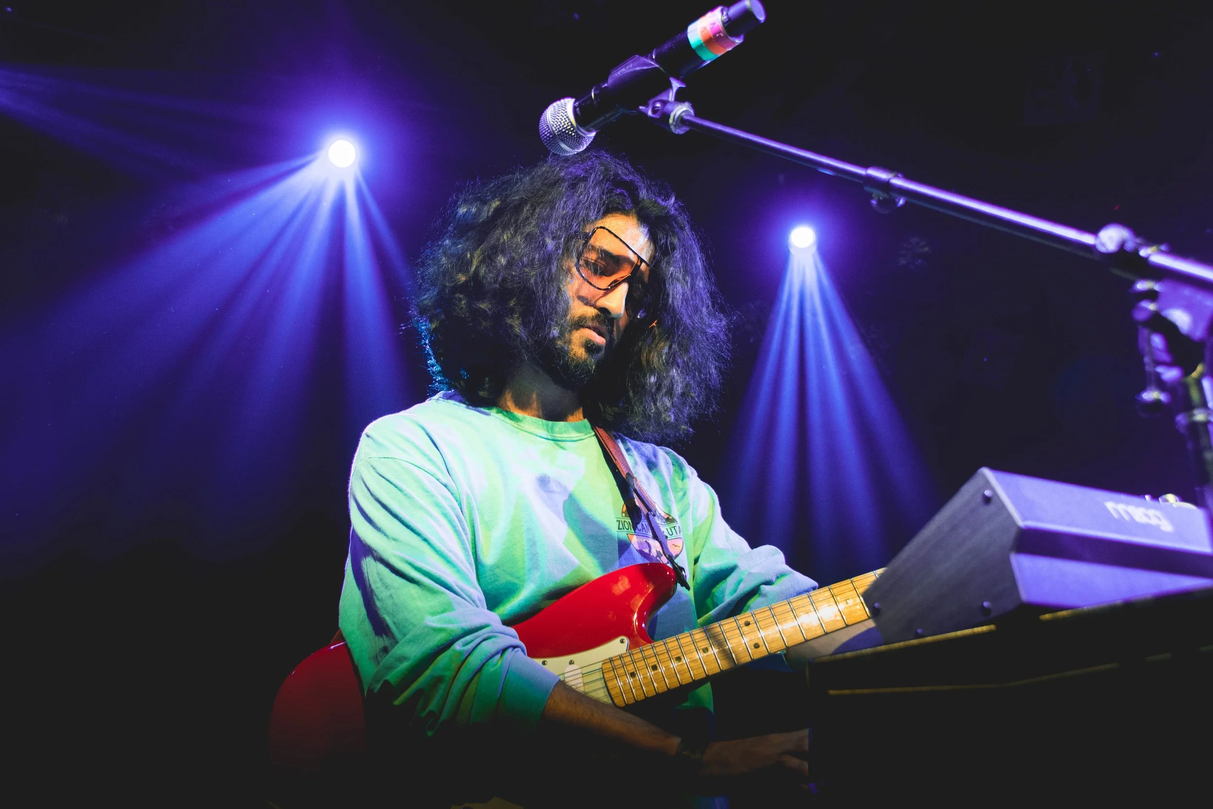 Joven músico tocando guitarra eléctrica y teclado en un escenario con luces azules y moradas.