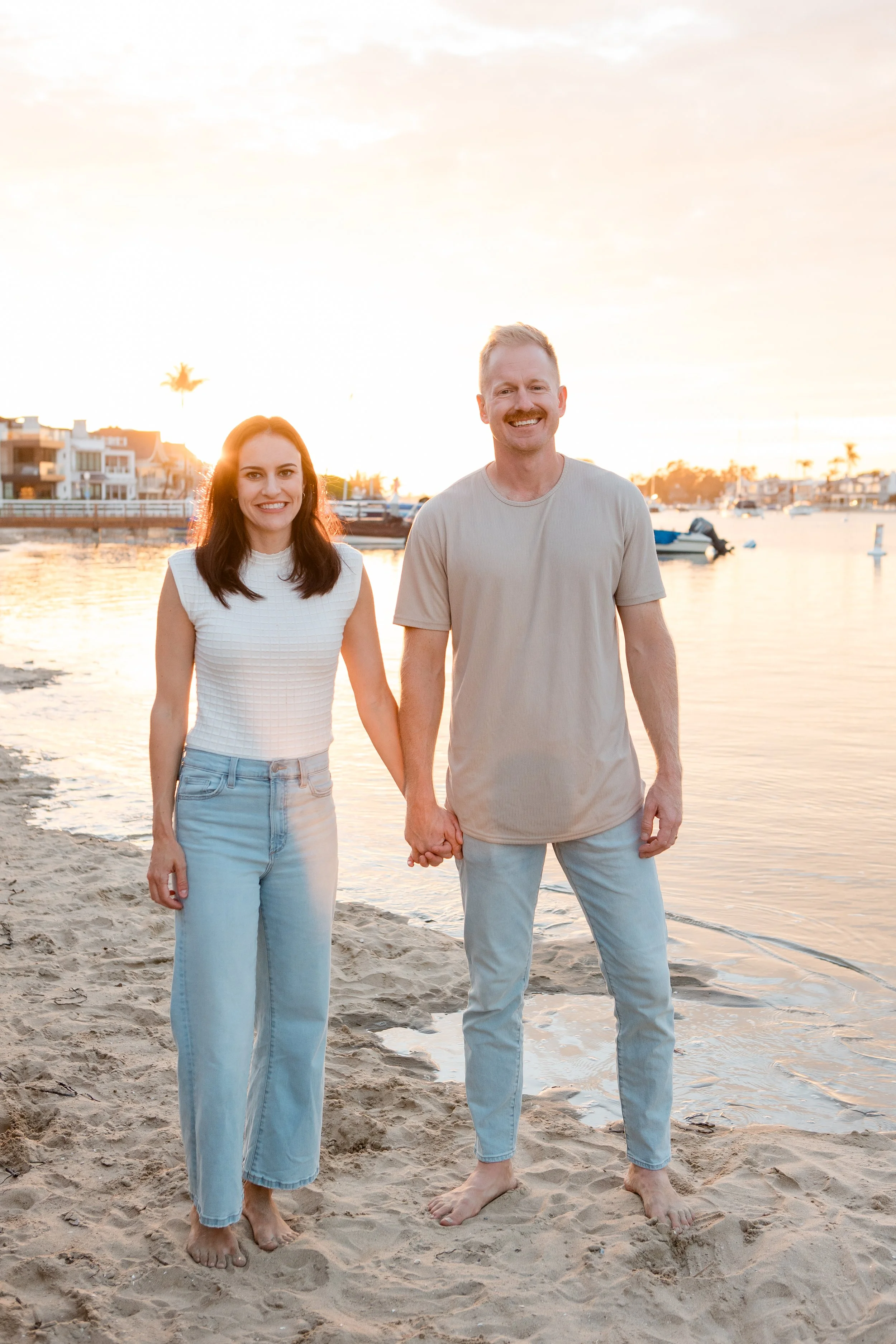 A smiling couple holding hands on a sandy beach at sunset, with boats and waterfront houses in the background.