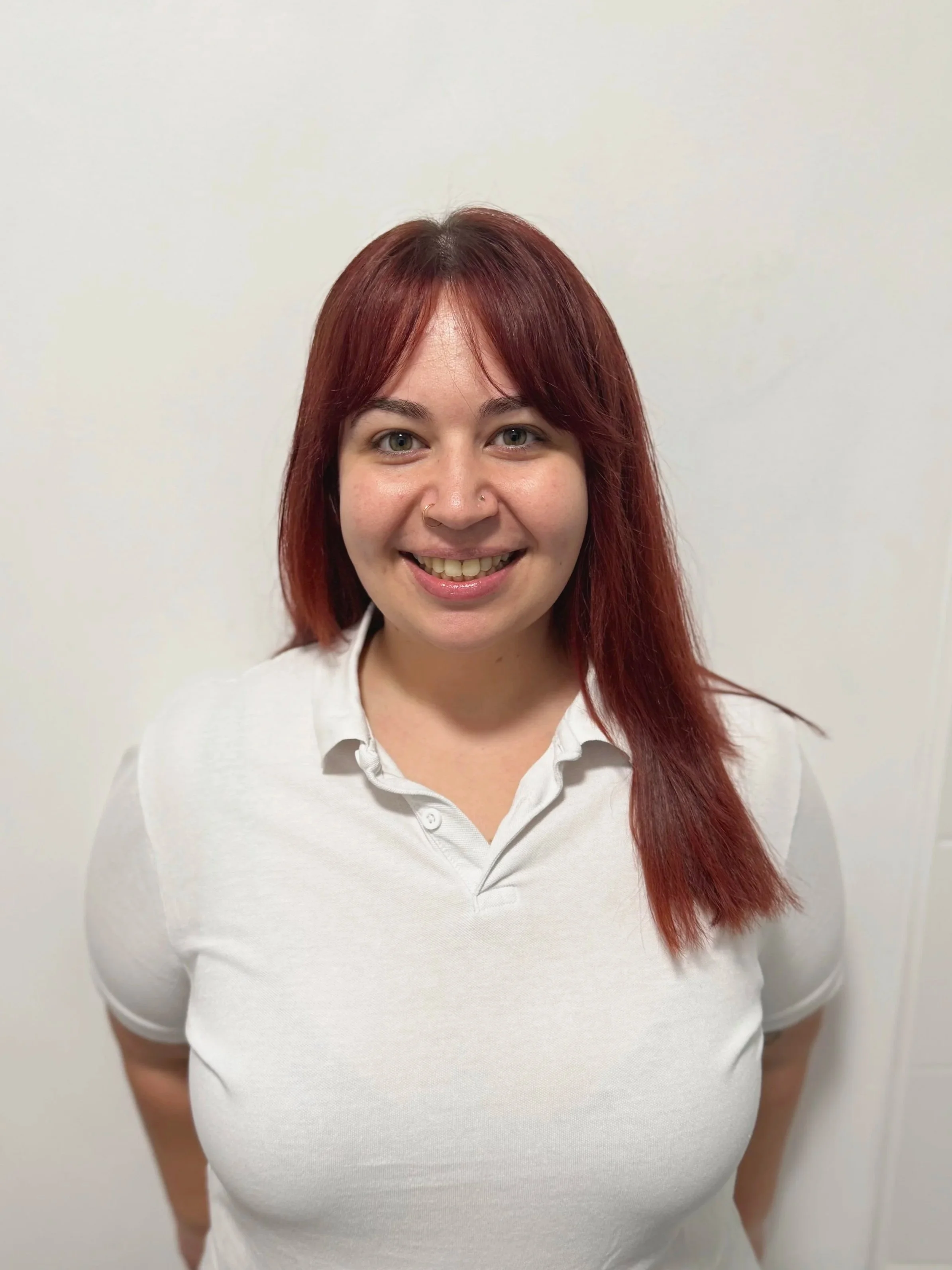 Young woman with red hair and fair skin smiling, wearing a white polo shirt, standing against a plain white wall.