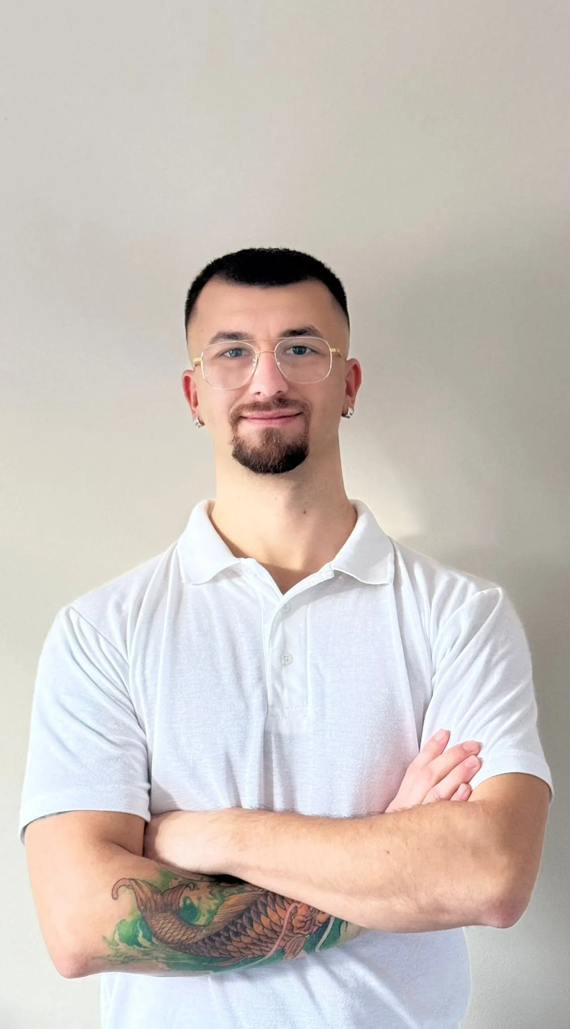 A young man with short black hair, glasses, and earrings, wearing a white polo shirt with his arms crossed, standing against a plain light-colored wall.