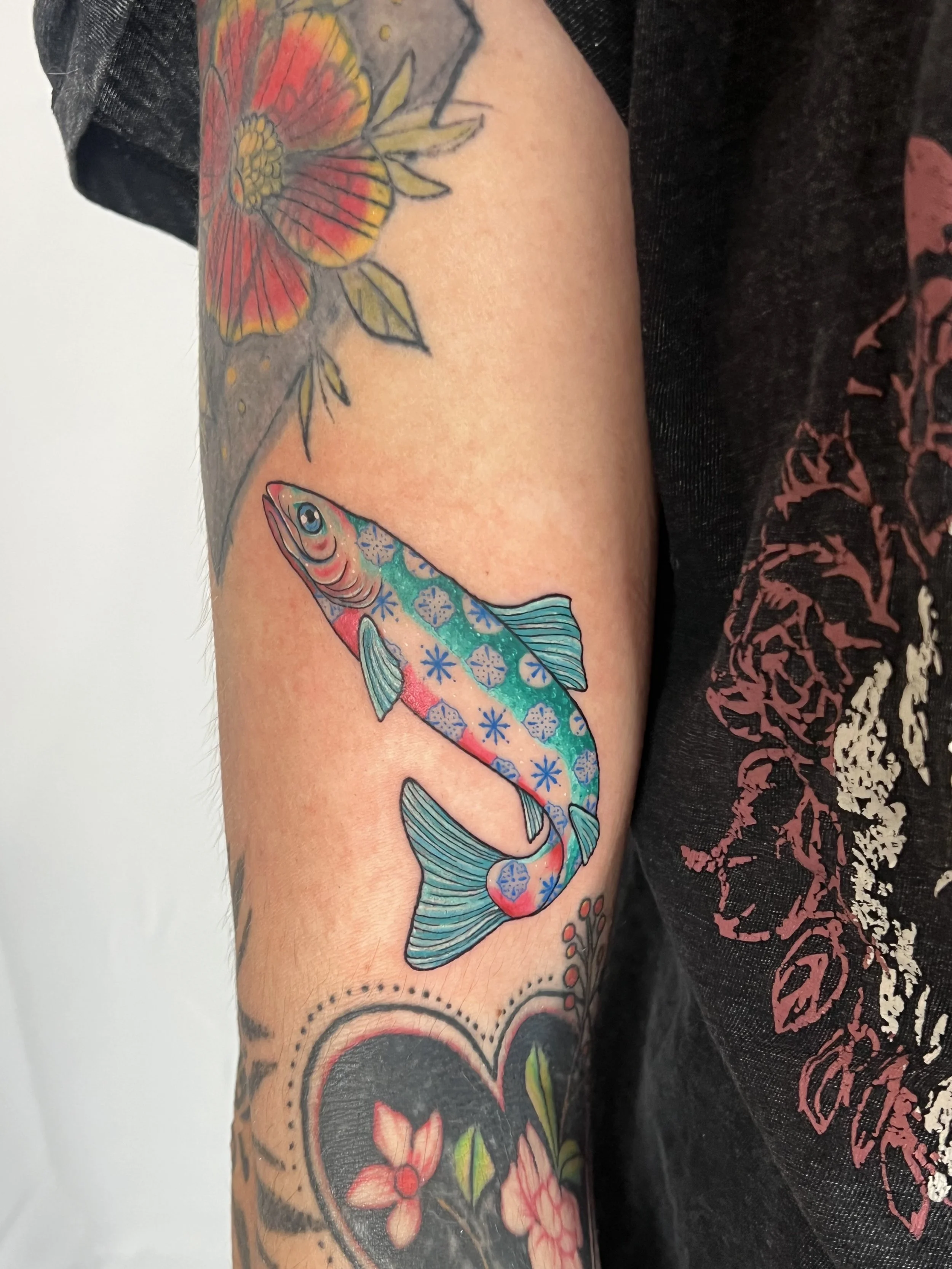 A person's arm with a colorful tattoo of a fish and a flower, along with additional tattoo designs on the arm.