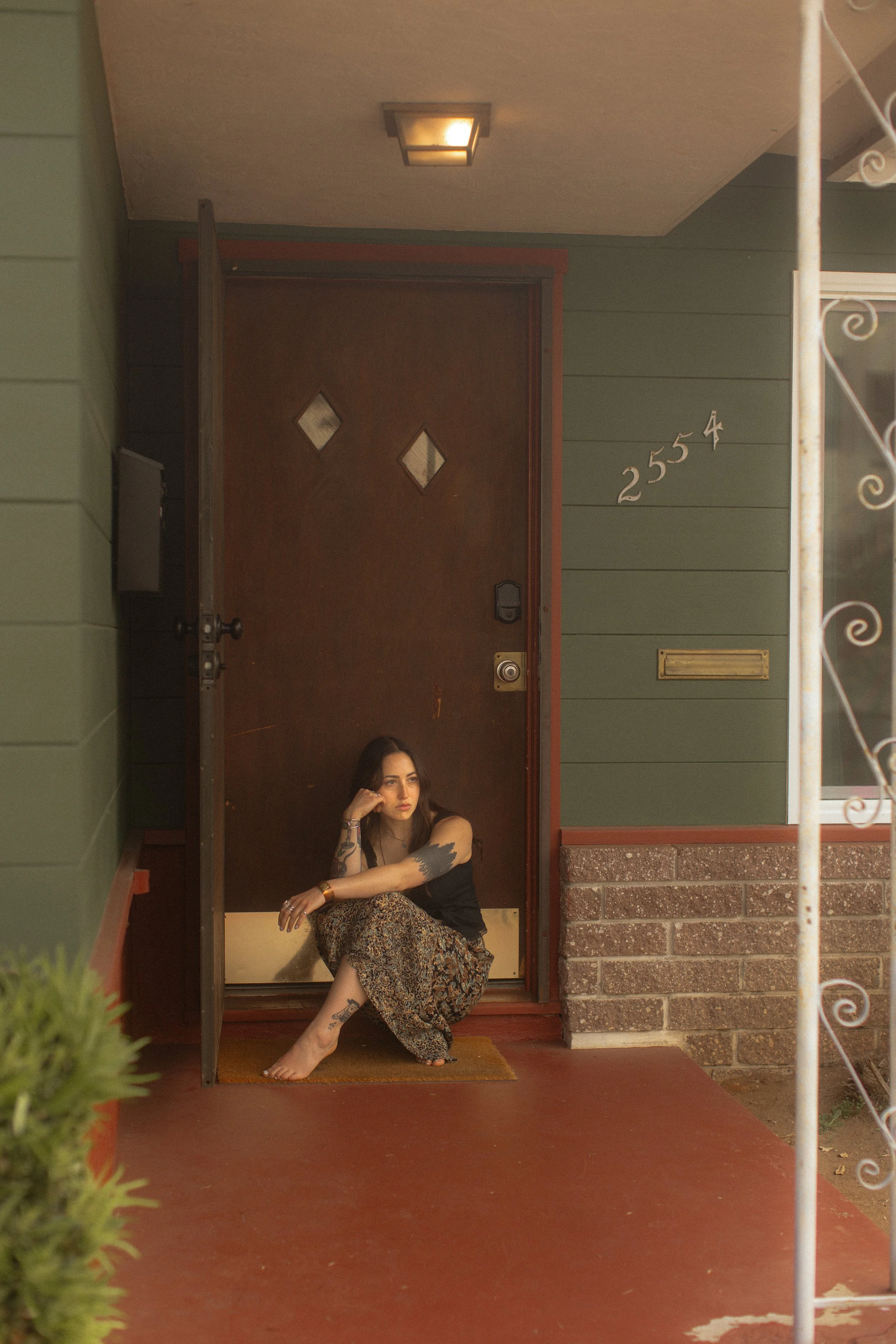 A woman with tattoos sitting on the porch of a house, looking off to the side. She is wearing a black top and patterned skirt, with one arm resting on her knee and her chin resting on her hand. The house has a green wall, a brown door with diamond-shaped windows, and a brick foundation. The house number "25554" is visible on the wall next to the door.