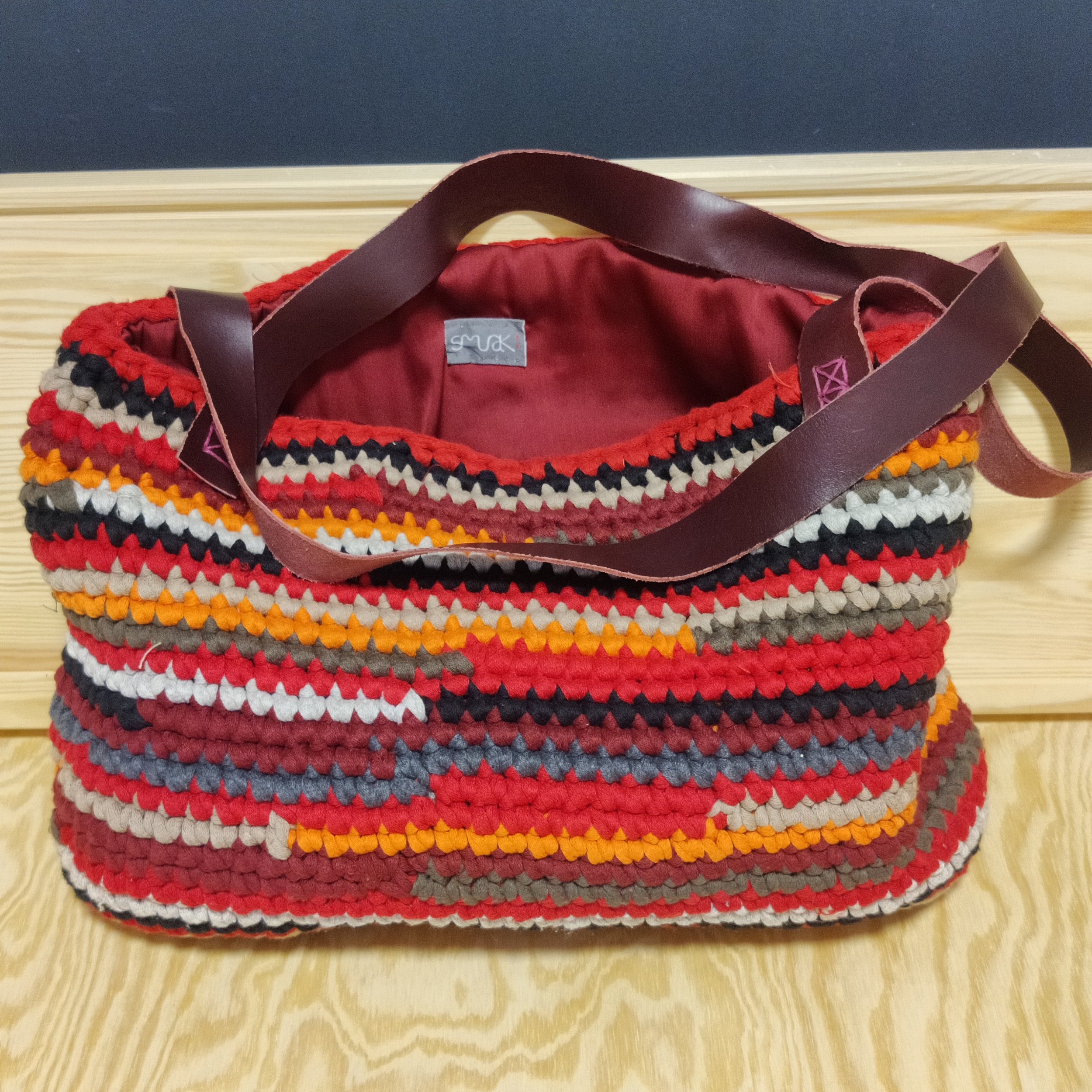 Bolso grande tejido con hilos de colores rojos, grises, blancos, negros y naranjas, con asas de cuero granate.