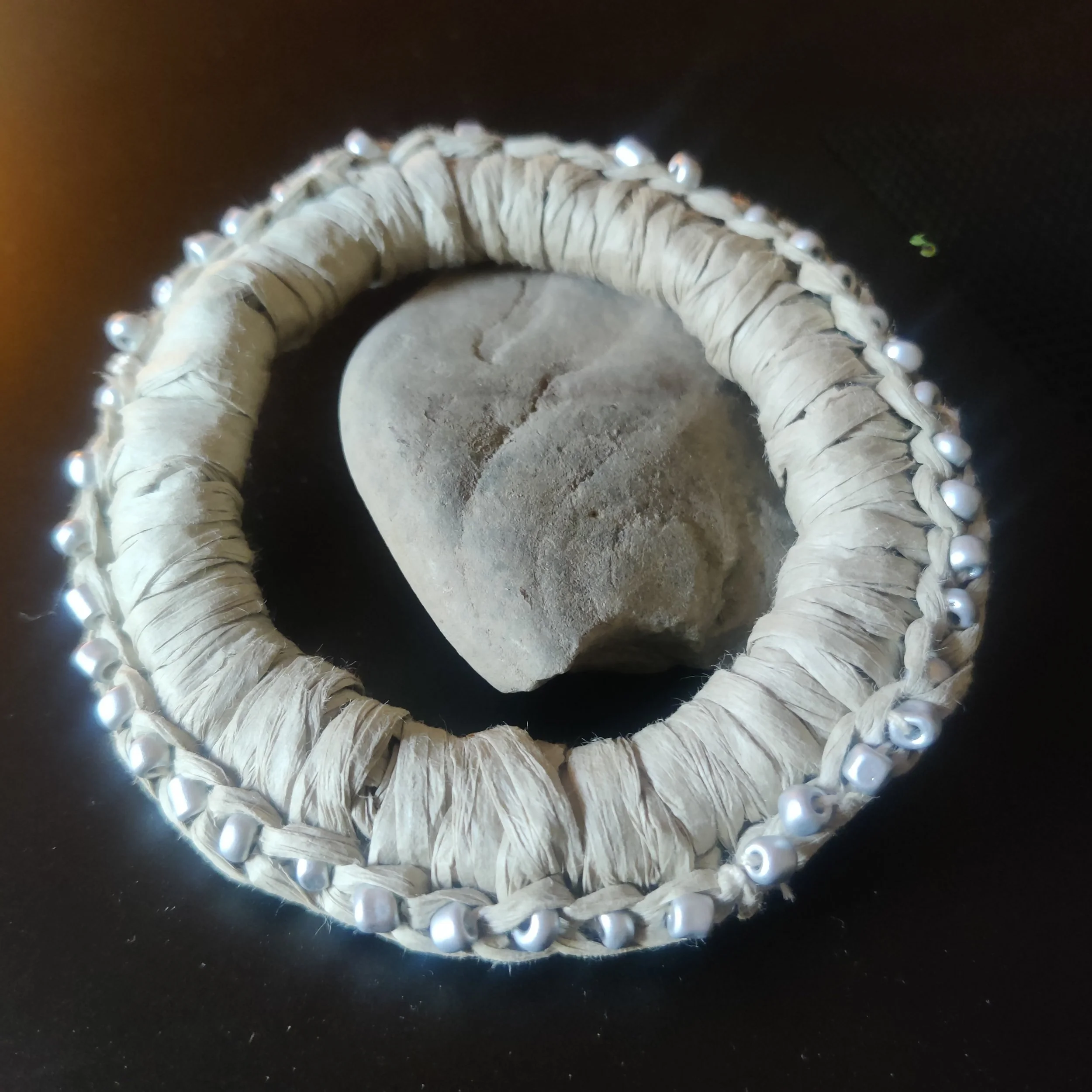 Pulsera decorativa con perlas y tela, colocada sobre una piedra en un fondo oscuro.